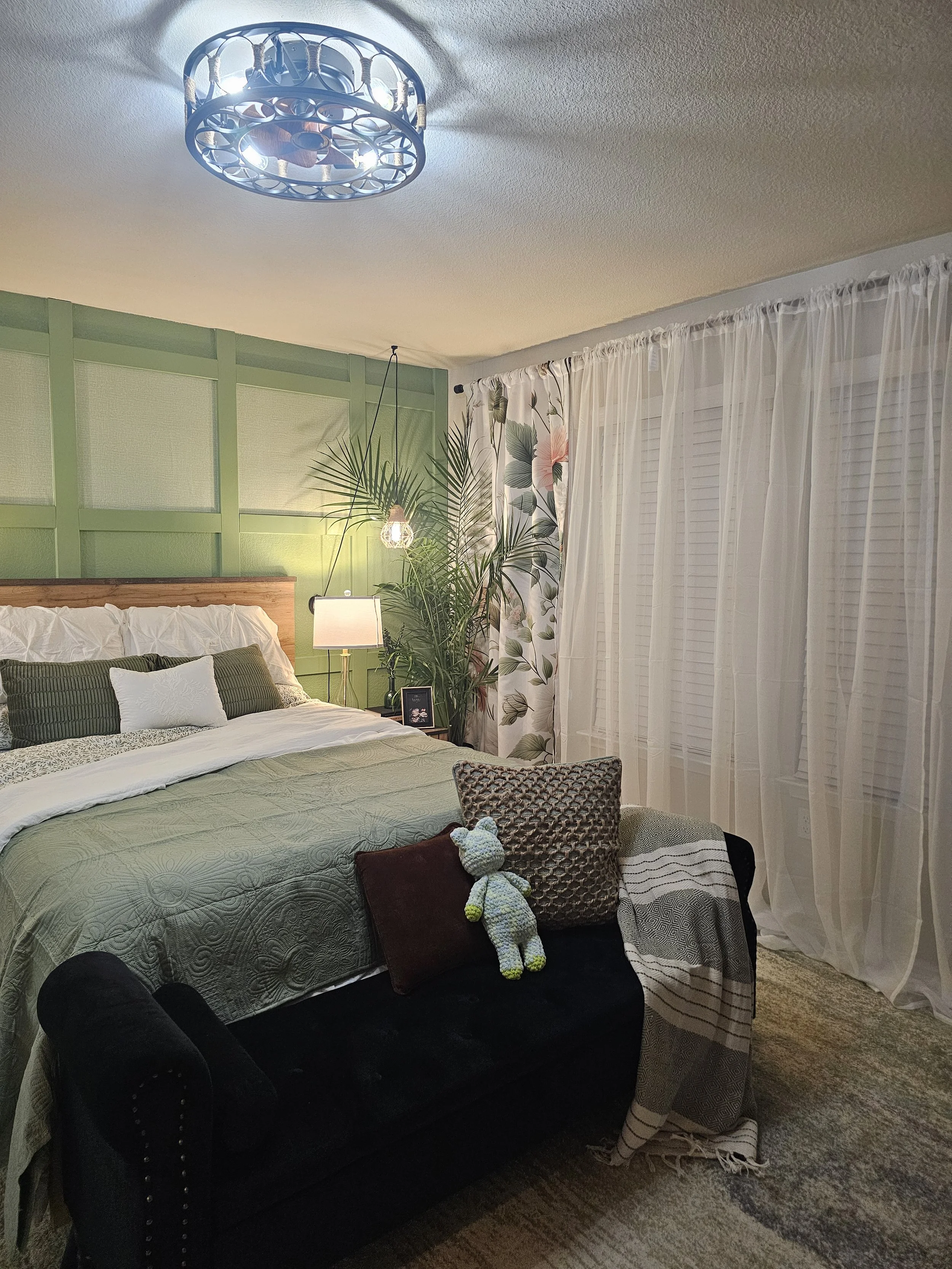 A bedroom with a bed, green wall behind headboard, potted plant, nightstand with lamp, framed photo, sheer curtains, and a black couch with pillows and stuffed animal.