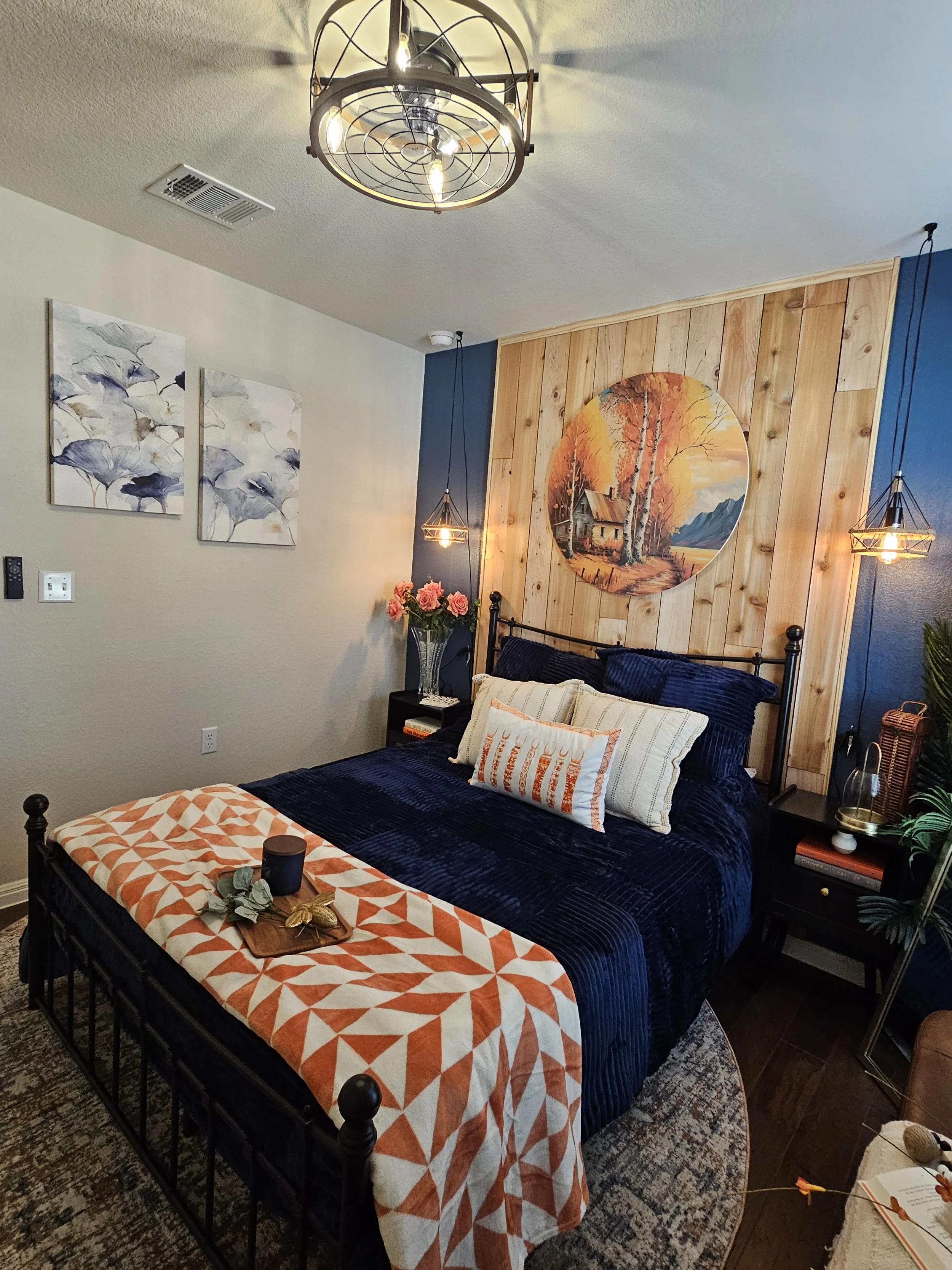 A bedroom with a black metal bed frame, navy blue bedding, and decorative pillows. The wall behind the bed has a wooden panel wall with a round landscape painting of a house, trees, and mountains. On the side, there is a black nightstand with books, a glass vase with pink roses, and a lamp. Two hanging pendant lights with a wireframe design provide illumination. The ceiling has a modern chandelier with exposed bulbs, and an air vent is visible. The bed has a white and orange geometric patterned throw blanket at the foot, with a small wooden tray holding decorations. The floor has a patterned rug.