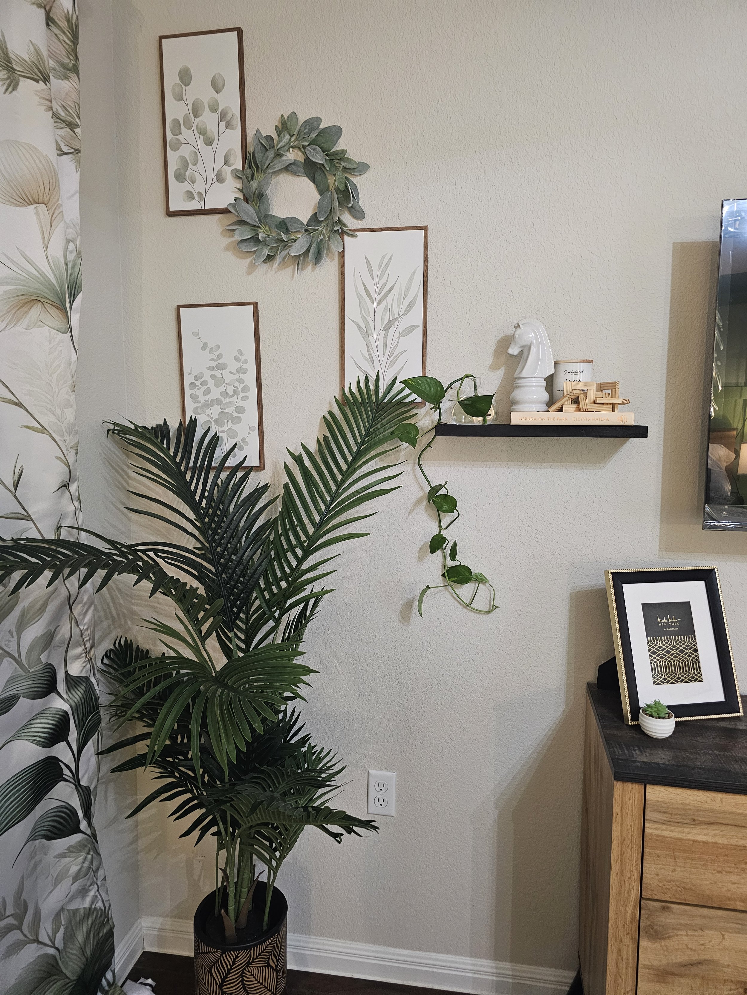 Living room corner with a large potted palm plant, framed botanical art on the wall, a black shelf with a white horse head sculpture, a small plant, and stacking books, a picture frame, and a small succulent on a wooden cabinet.