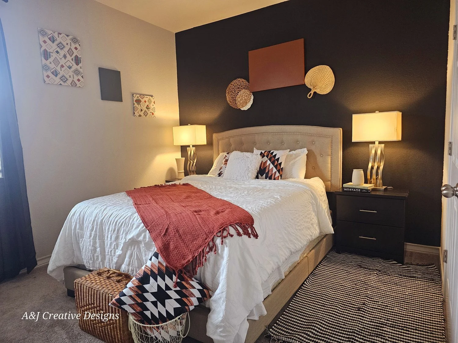 A decorated bedroom with a beige upholstered bed, white bedding, and decorative pillows. There are two nightstands with modern lamps, a black and white rug, abstract wall art, and a dark accent wall.