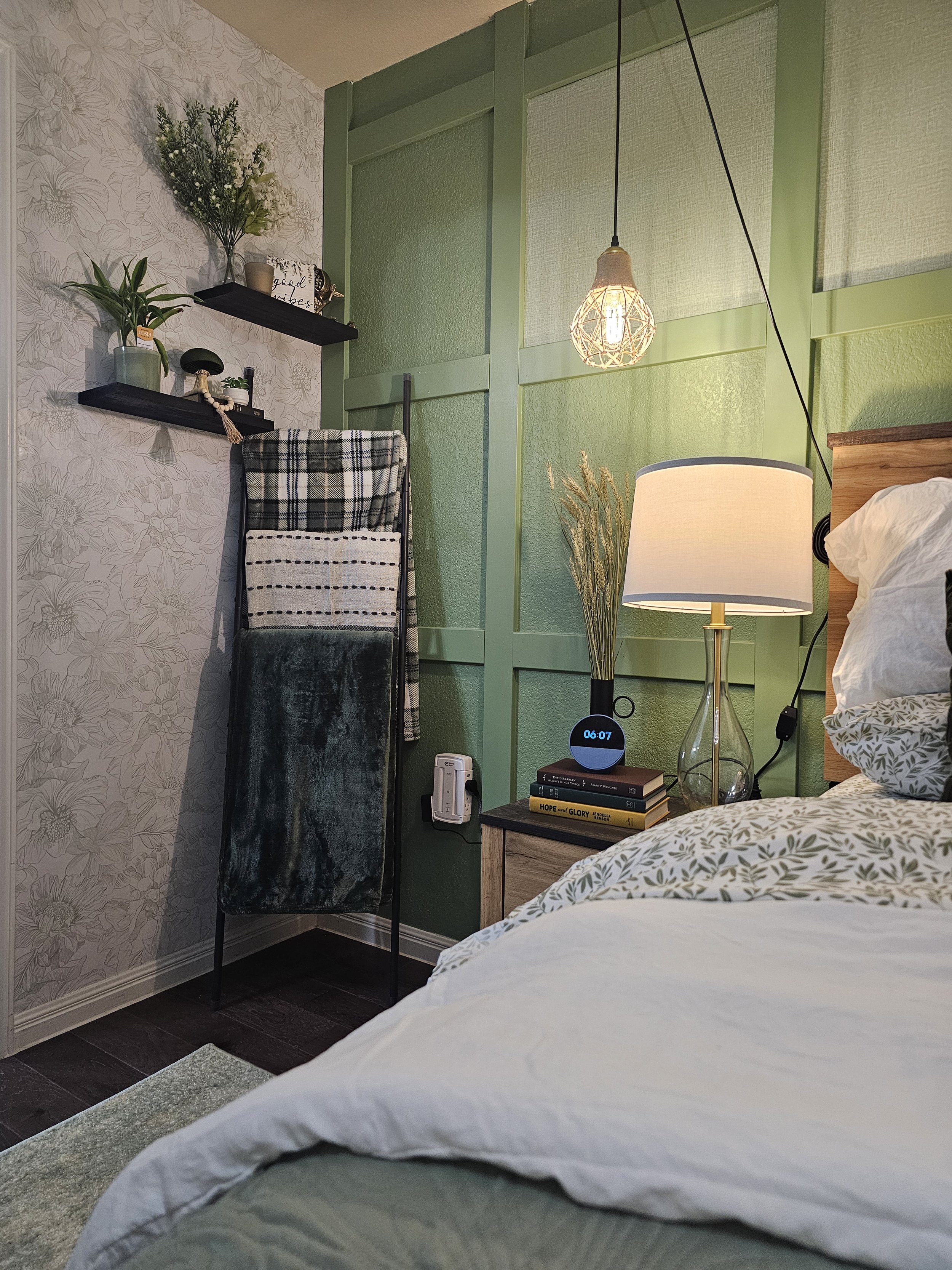 A bedroom corner with a green wall, a bed with white and floral bedding, a wooden nightstand with a table lamp, books, and a digital clock showing 6:07. An artful light fixture hangs from the ceiling. To the left, there is a wall with floral wallpape