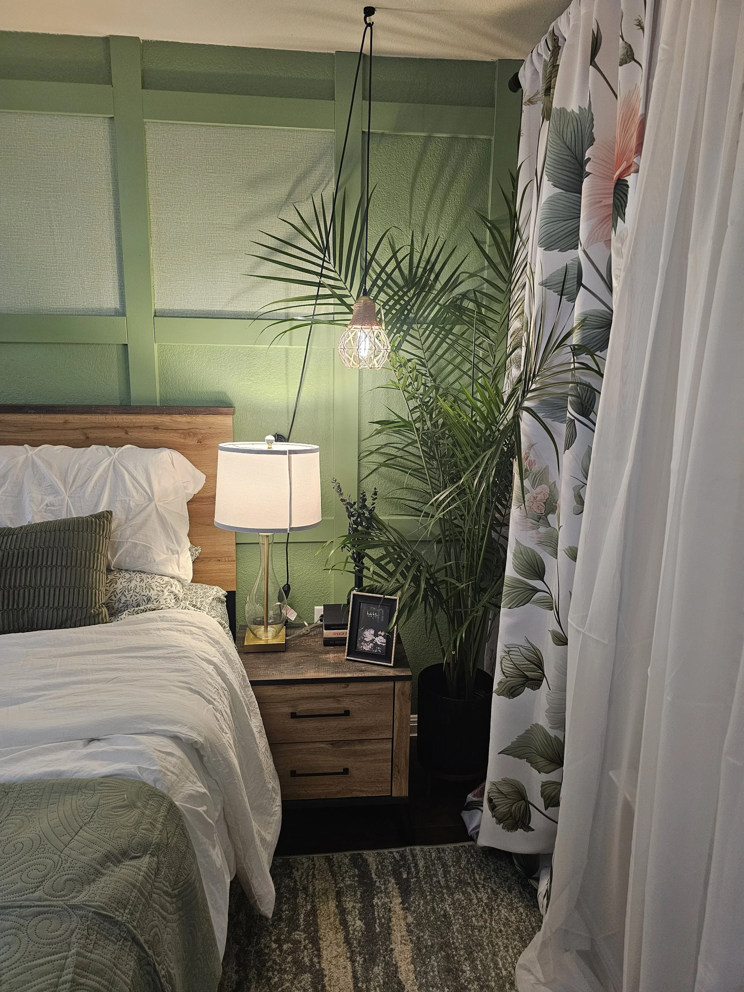 A bedroom corner with a bed, bedside table, lamp, large potted plant, and floral curtains.