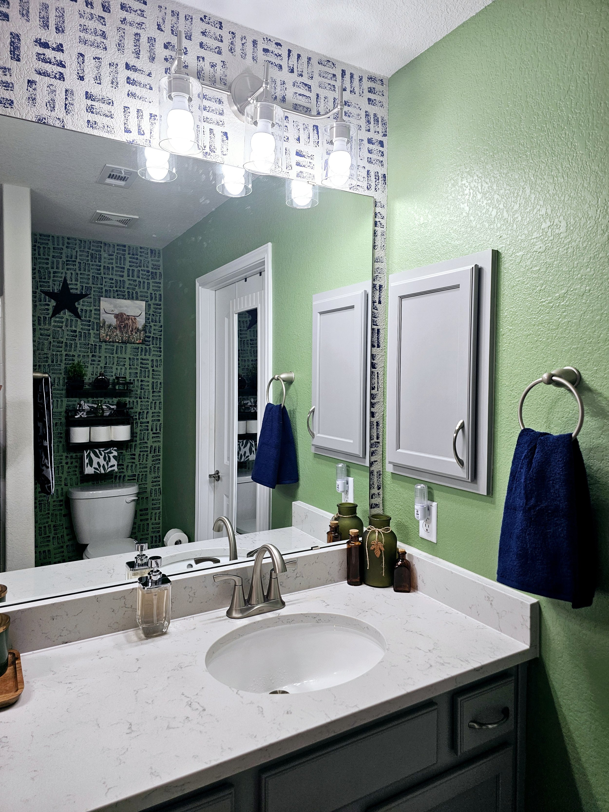 A modern bathroom with a white marble countertop, dark gray cabinet beneath, a large mirror, and a white sink. The counter has small bottles and decorative jars. Bright lighting is from a three-bulb fixture. The wall behind the mirror is painted green with textured finish. Towel rings with blue towels are on the sides, and a doorway leads to a small toilet area with dark patterned wallpaper, a picture of a highland cow, and a star-shaped decoration.