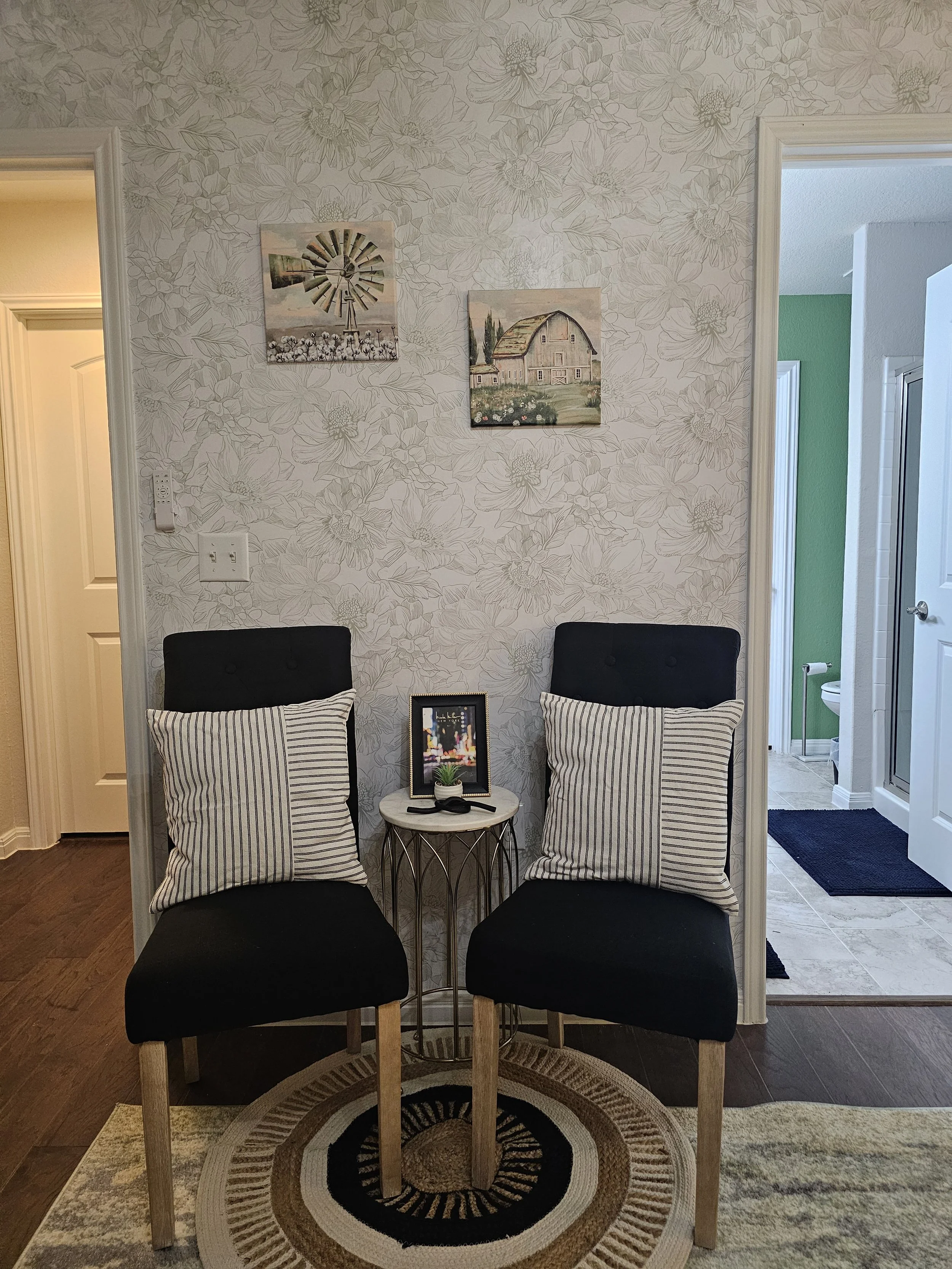 Two black upholstered chairs with striped pillows, a small round table with a framed photo, a small potted plant, and a remote control, set against a wall with floral wallpaper. Artwork decorates the wall, with doorways on both sides leading to diffe