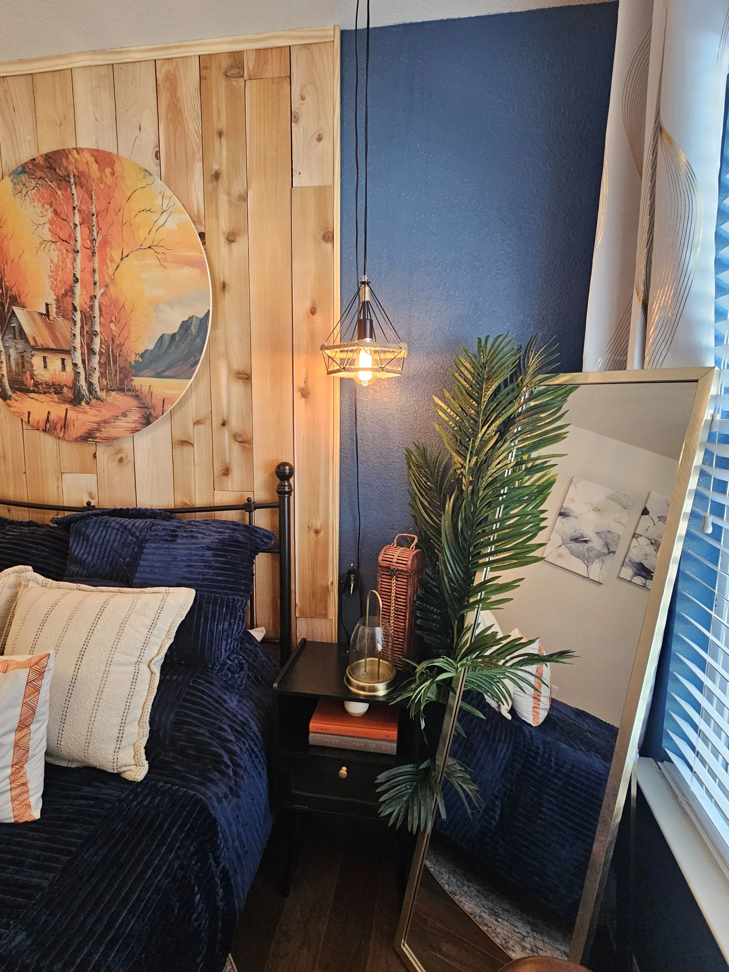 A bedroom with a wooden headboard, dark blue bedding, and decorative pillows. A black nightstand holds a glass with a gold base, a small basket, and a closed book. A large green potted plant is beside a mirror. A warm pendant light hangs from the ceiling. The wall behind the bed has a painting of autumn trees and mountains, and part of a window with white curtains and blinds is visible.