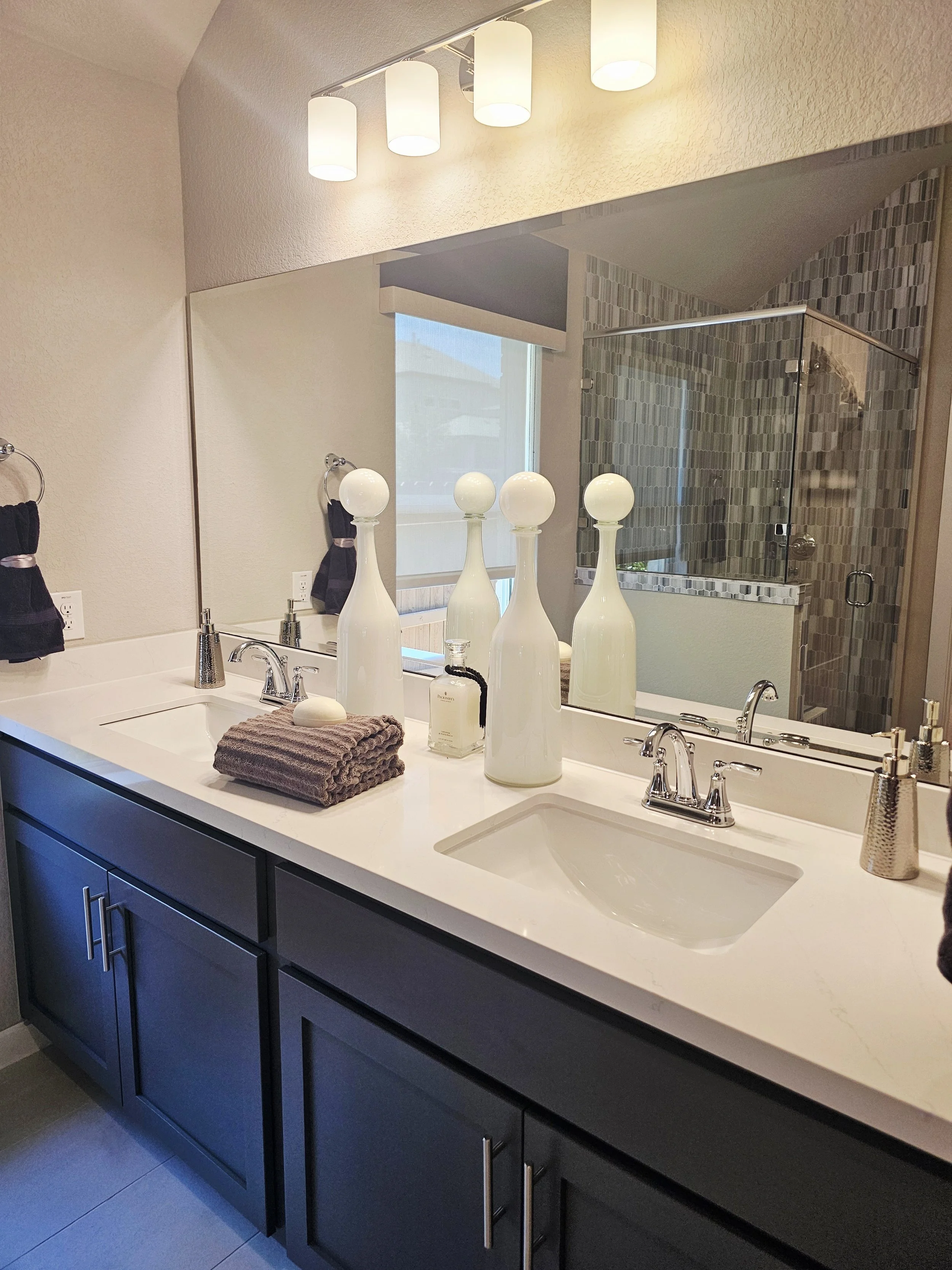 Modern bathroom vanity with a white countertop, a double sink, and dark blue cabinets. The countertop is decorated with four white vases, a soap dispenser, and a folded towel. A mirror spans the length of the vanity, and toilet paper is mounted on the wall. A window with a shade and a walk-in shower with a glass door and mosaic tile interior are visible in the reflection.