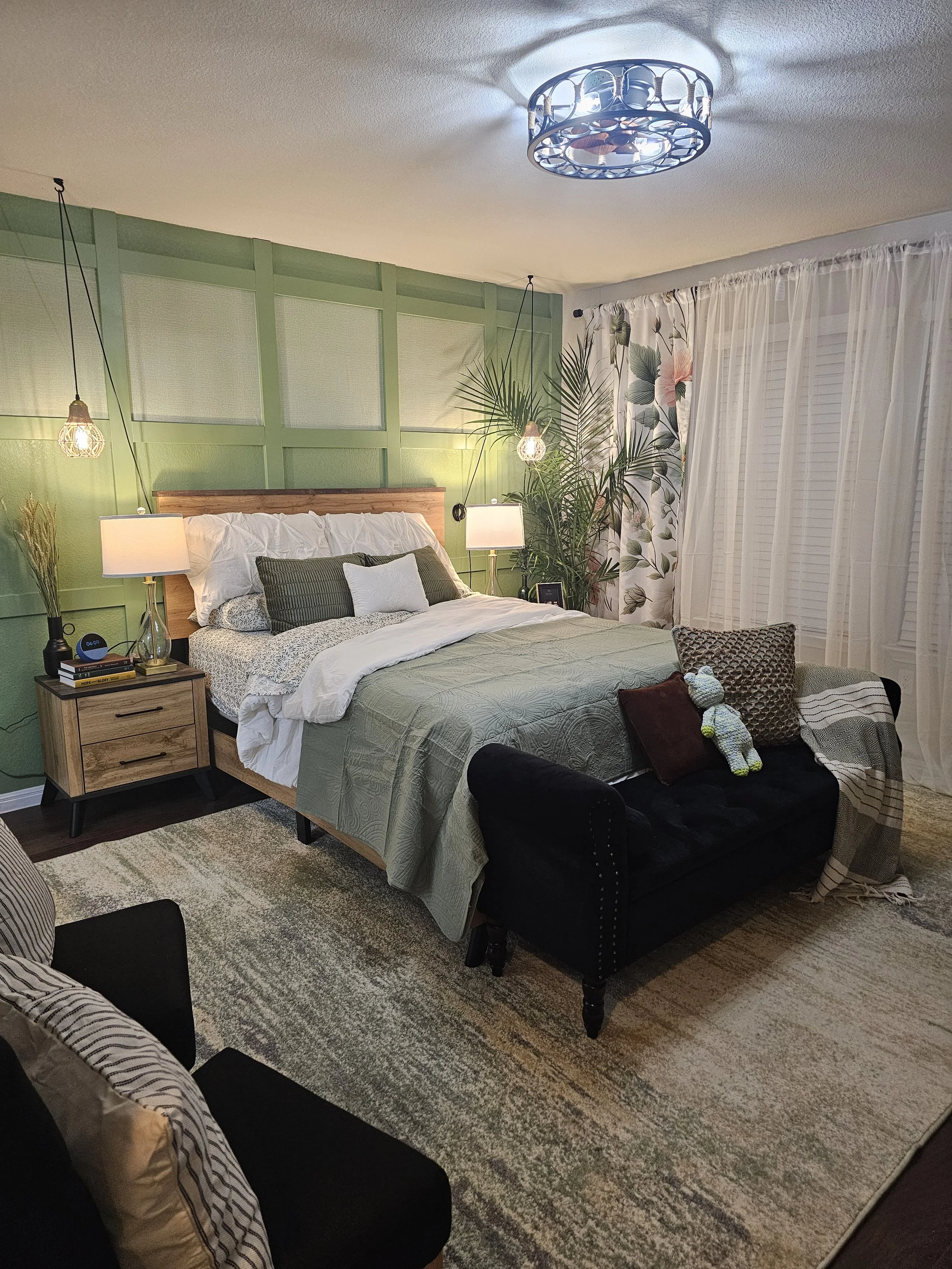 A cozy bedroom with a green accent wall, a bed with white and dark green pillows, and a wooden headboard. Two hanging pendant lights and bedside lamps illuminate the room. A black loveseat with decorative pillows and a blanket at the foot of the bed.