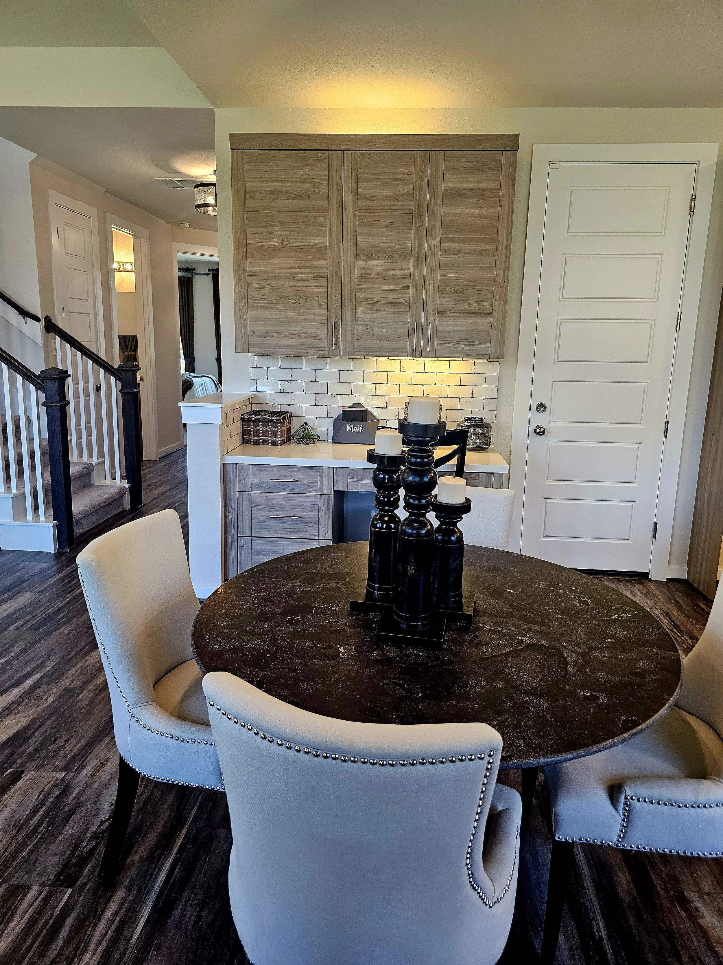 Dining room with a black round table, six beige upholstered chairs with silver nailhead trim, a dark wood floor, a white wall with a built-in wooden cabinet above a white brick backsplash, and a black candlestick centerpiece with white candles.