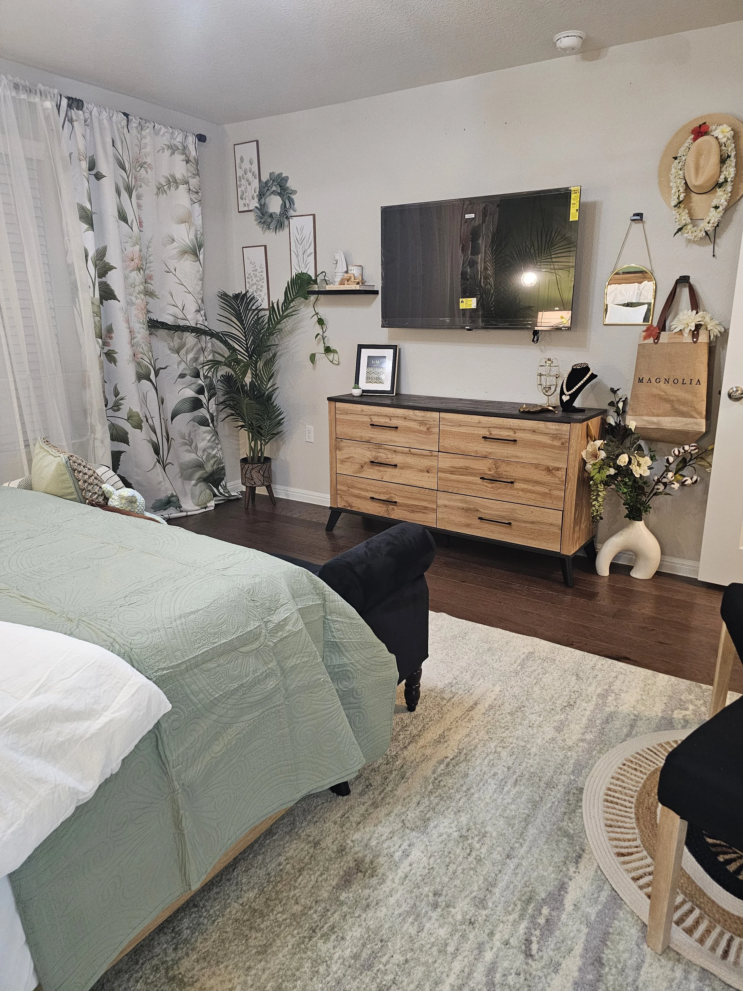Living room with a green quilted sofa, a potted plant near the window with floral curtains, a wooden dresser with a mounted TV, decorated with flowers, hats, mirrors, and framed artwork, and a patterned area rug on dark hardwood flooring.