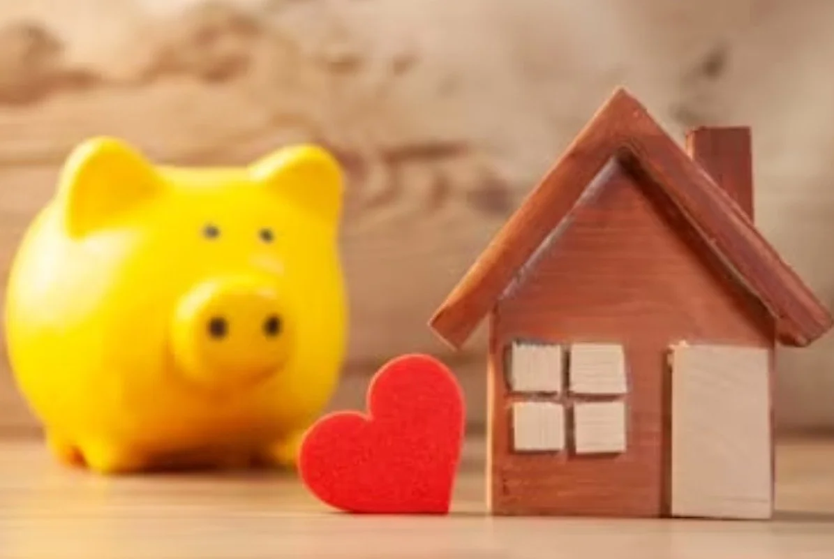 Yellow piggy bank, red heart, and small wooden house figurine on a wooden surface.