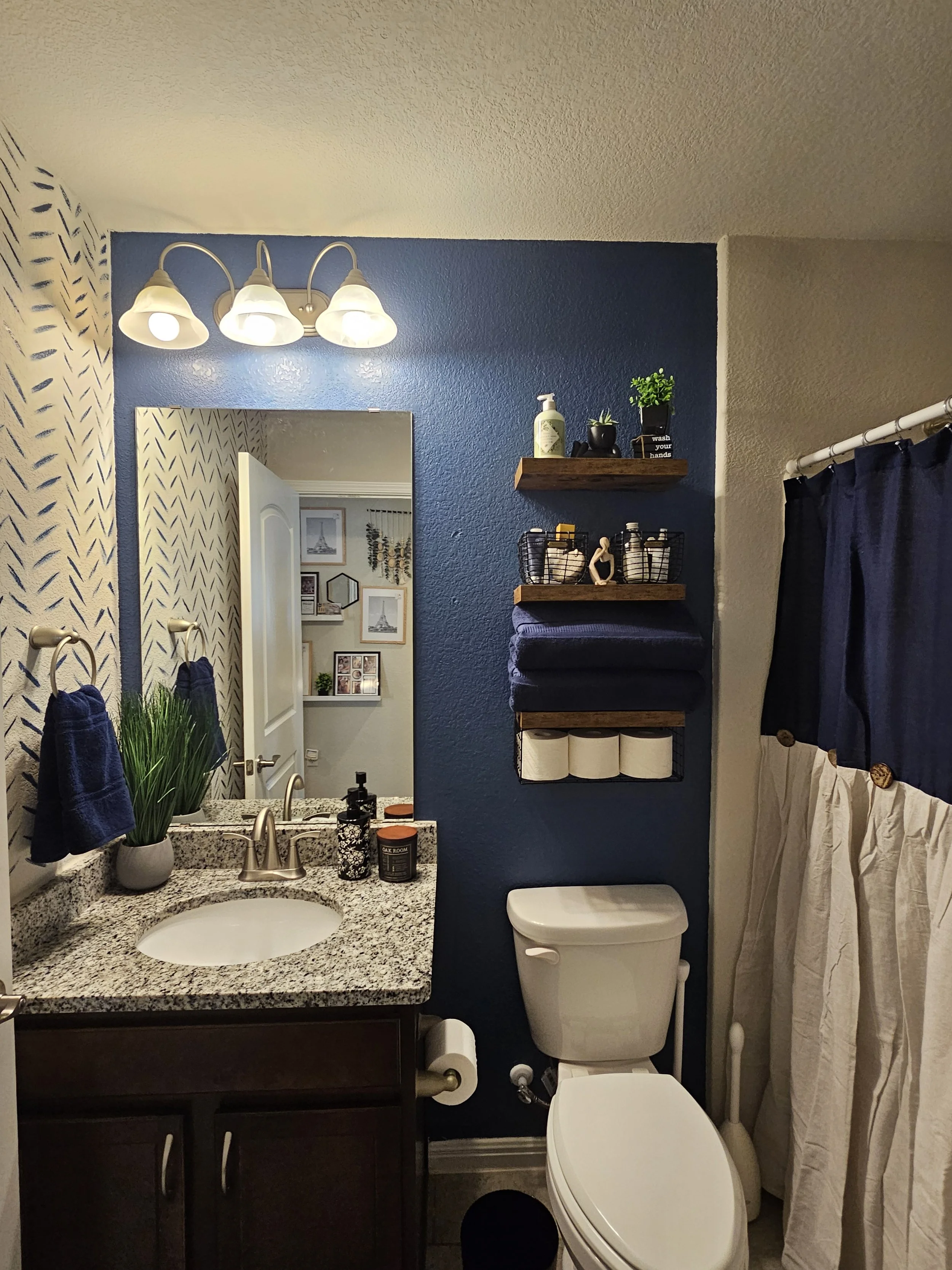 Bathroom with blue accent wall, granite countertop vanity with oval sink, potted plant, soap dispenser, towel rings with blue towels, framed pictures and decor in mirror reflection, toilet with a black mat, wall shelves with plants, toiletries, and toilet paper, dark curtains, and a white curtain covering a window or shower.