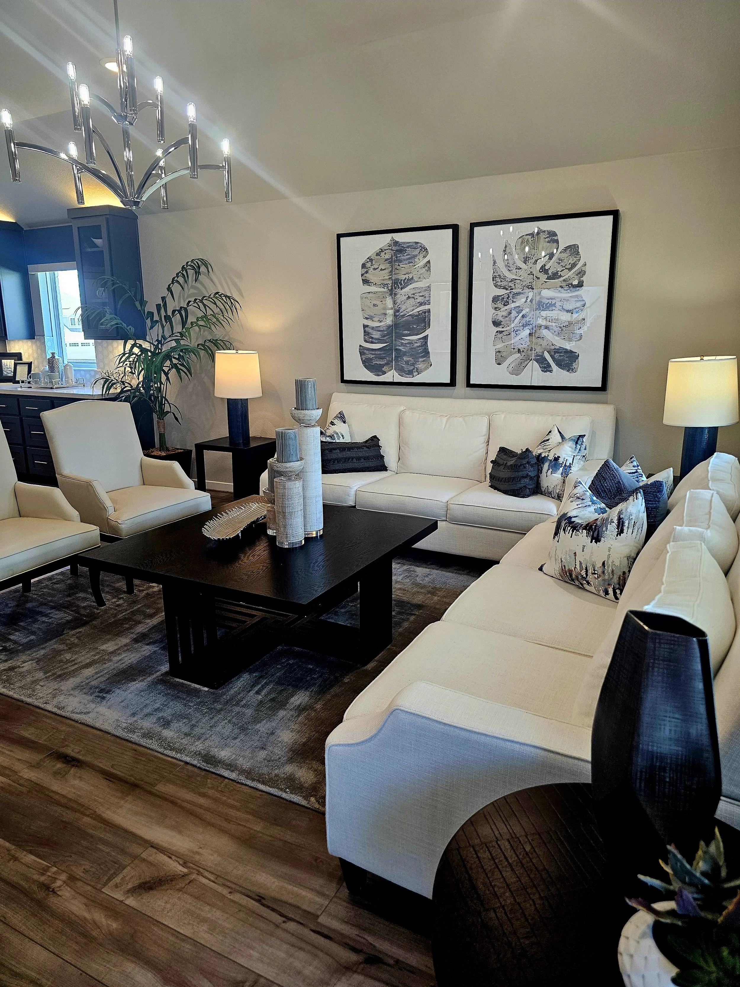 A living room with beige walls, a white sectional sofa with black and white throw pillows, a black coffee table with decorative vases, two black table lamps with beige shades, two framed abstract art pieces of dark leaf patterns, a tall potted plant, a chandelier with multiple light bulbs, and a wooden floor with a patterned rug.