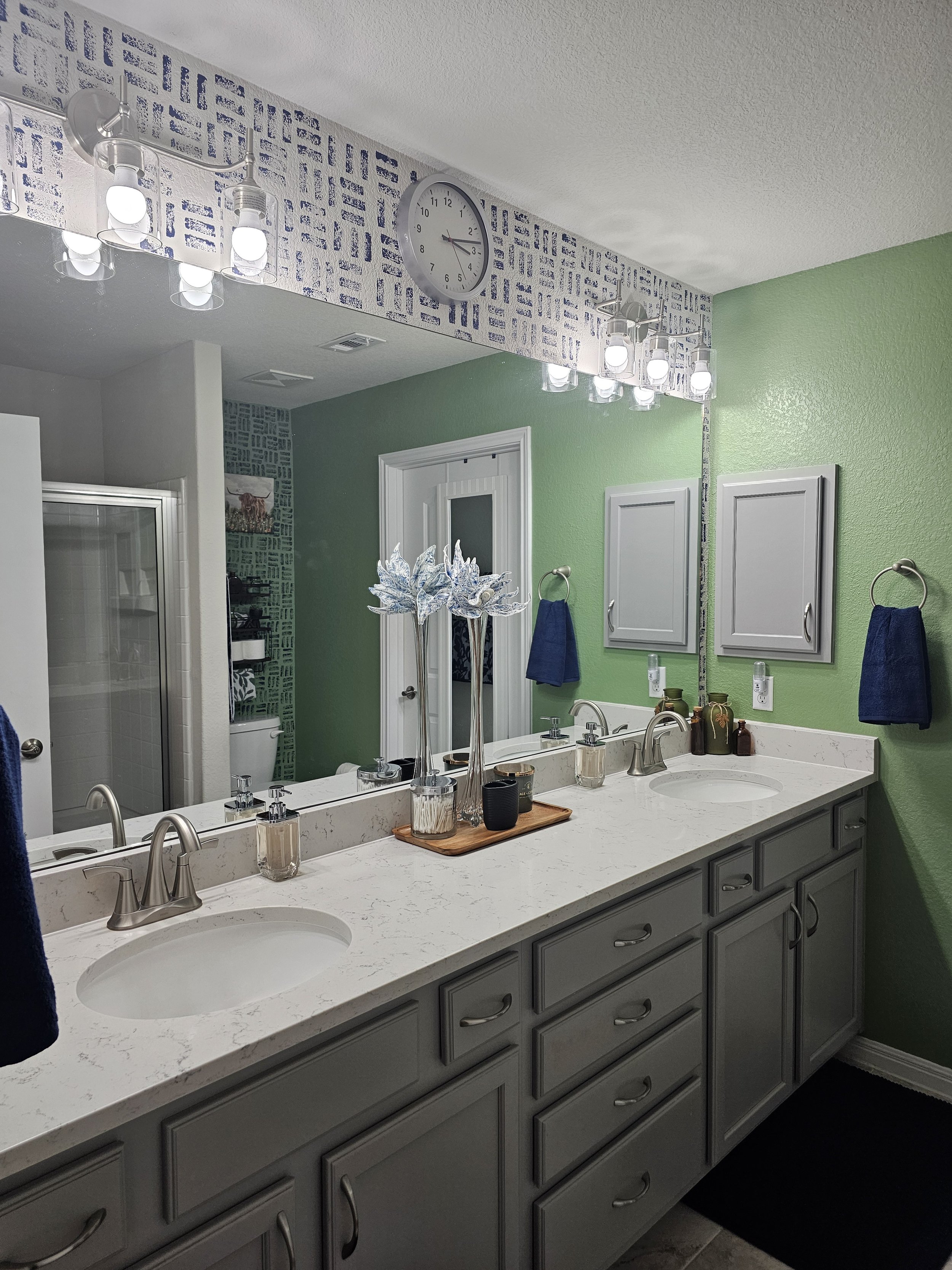 Bathroom with green wall, large mirror, double sink, gray cabinets, blue towels, wall art, decorative vases, and modern light fixtures.