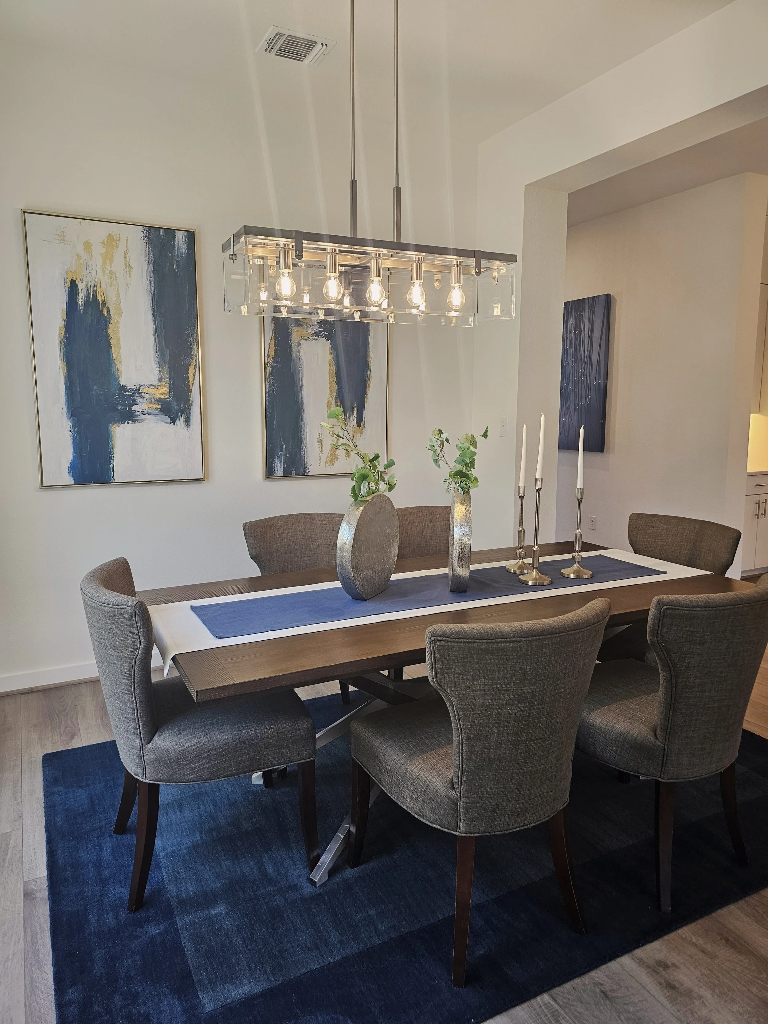 A dining room with a rectangular wooden table surrounded by six upholstered chairs, a blue and white table runner, three tall silver candlesticks with white candles, two vases with green foliage, a modern light fixture with exposed bulbs hanging from the ceiling, abstract blue, white, and gold artwork on the walls, and a dark blue rug under the table.