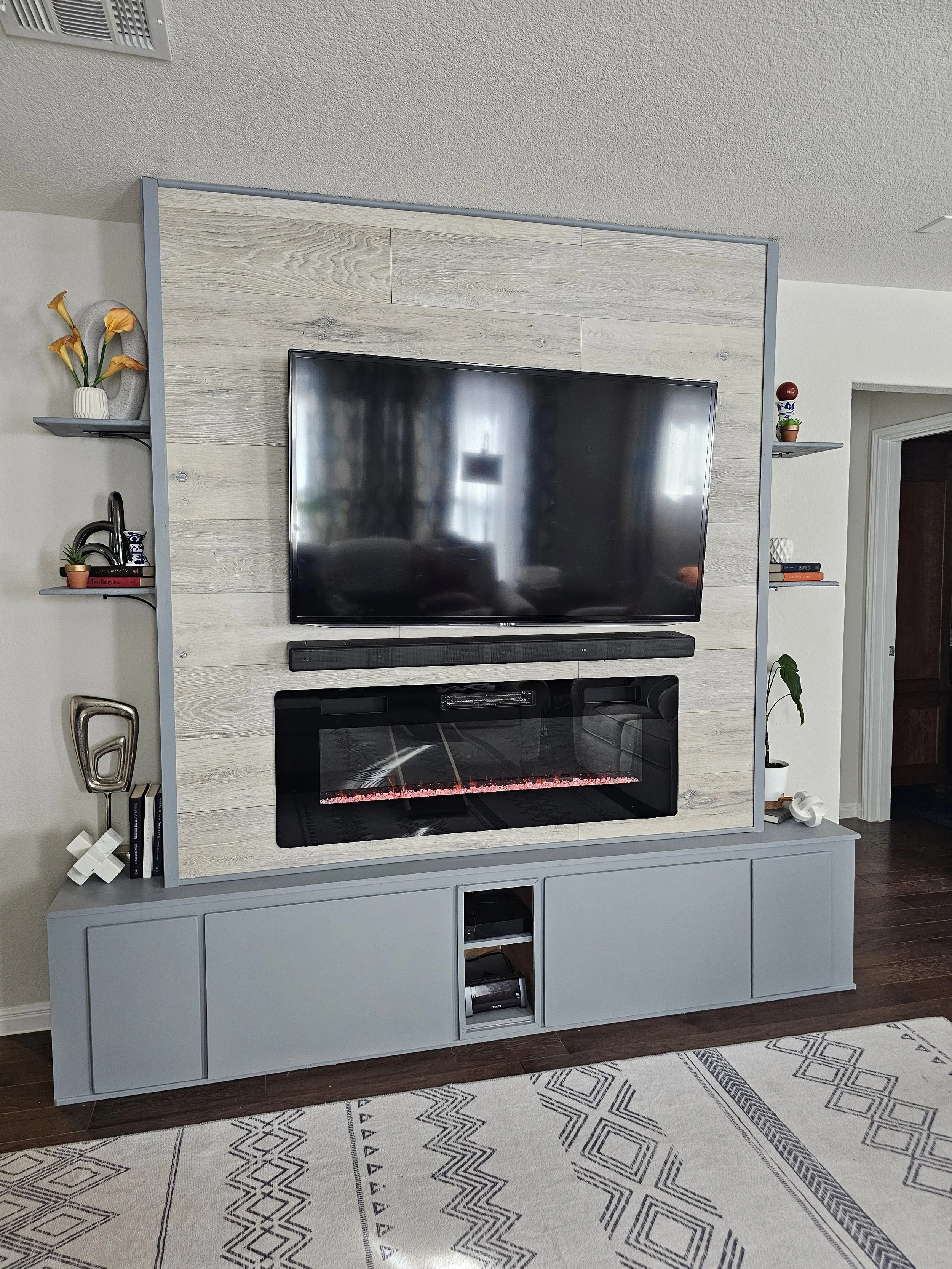 Living room entertainment center with a flat-screen TV, electric fireplace, and decorative items on shelves, with a patterned rug on a hardwood floor.
