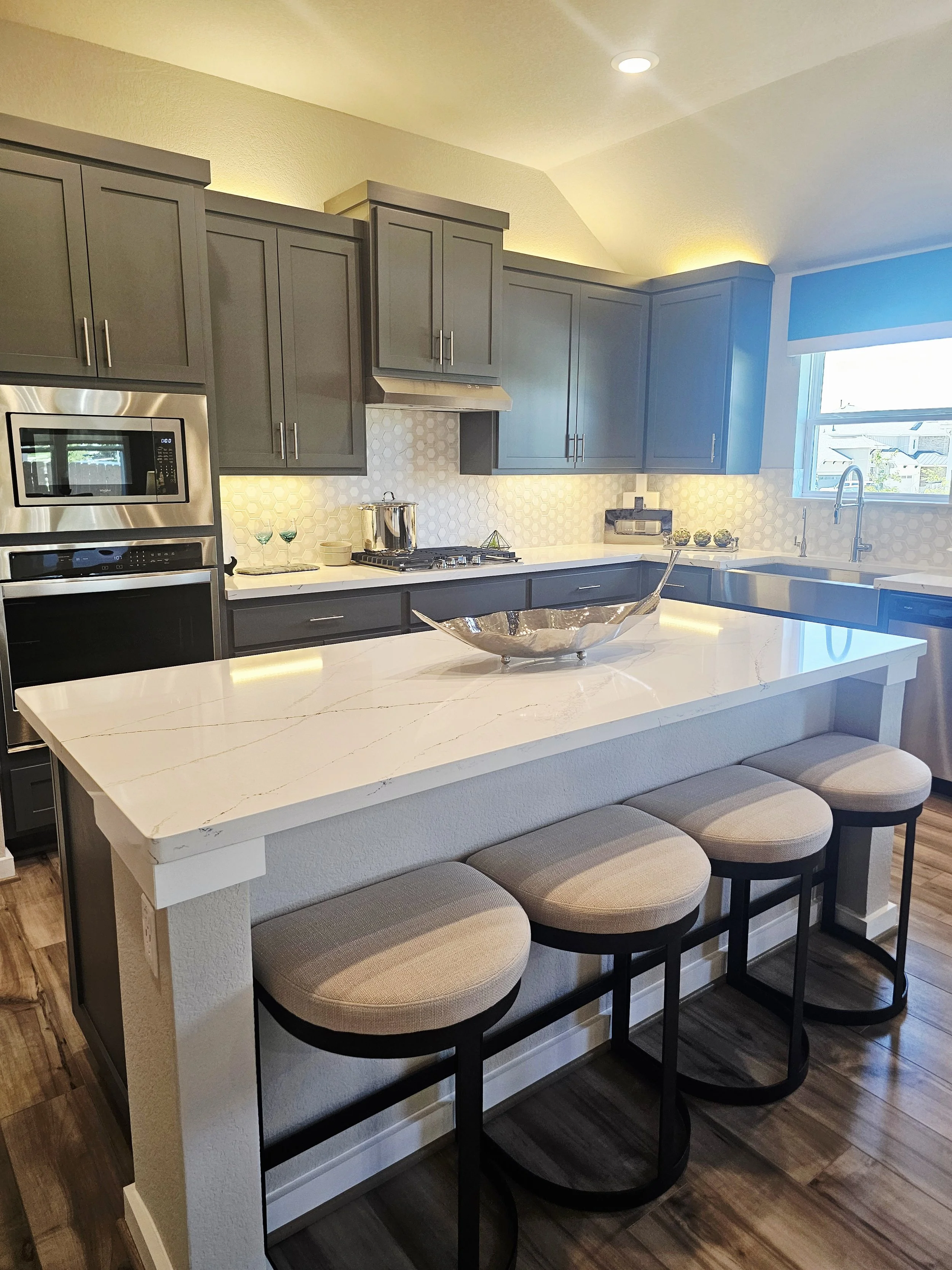 Modern kitchen with gray cabinets, white marble island, and four upholstered bar stools with black frames, wooden flooring, and a window with blue blinds.