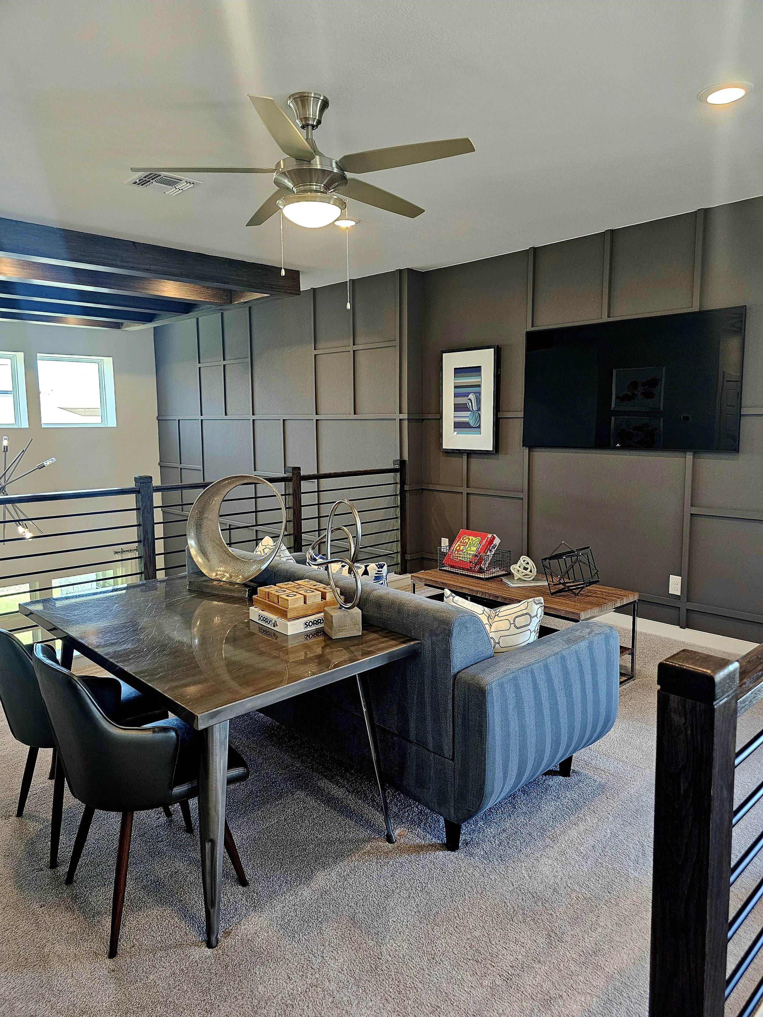 A modern living room with a wood dining table, black chairs, a gray striped sofa, and a wooden coffee table. The wall has gray paneling, a framed artwork, and a large flat-screen TV. There's a ceiling fan and small windows.