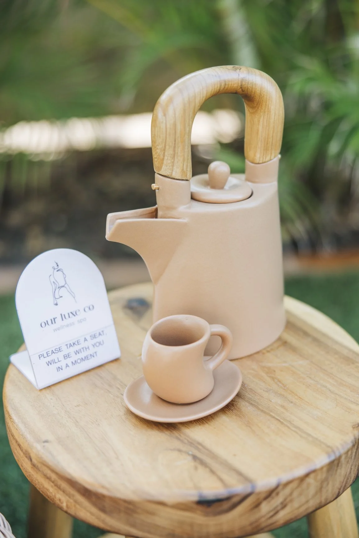 A beige ceramic teapot with a wooden handle on a wooden table, with a matching small cup and saucer, and a white sign that reads 'please take a seat, will be with you in a moment' in front of a blurred outdoor garden background.