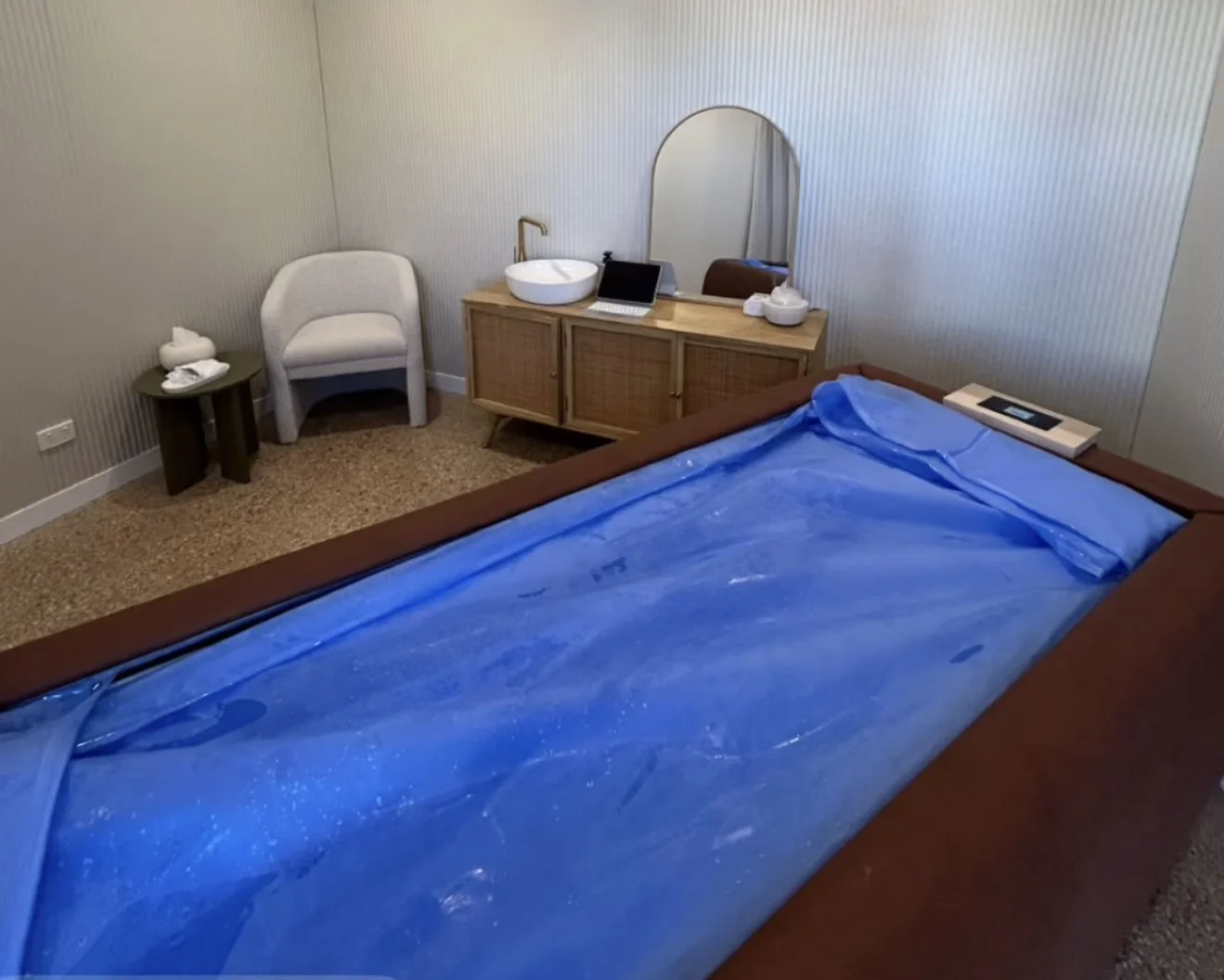 A bedroom with a bed covered in blue plastic, a nightstand with tissue box, a chair, and a wooden dresser with a sink and mirror on top.