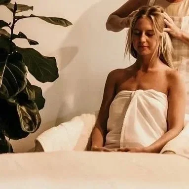 A woman giving a forehead massage and a hand on her shoulder to a woman sitting with her eyes closed, wrapped in a towel, in a massage or therapy room.