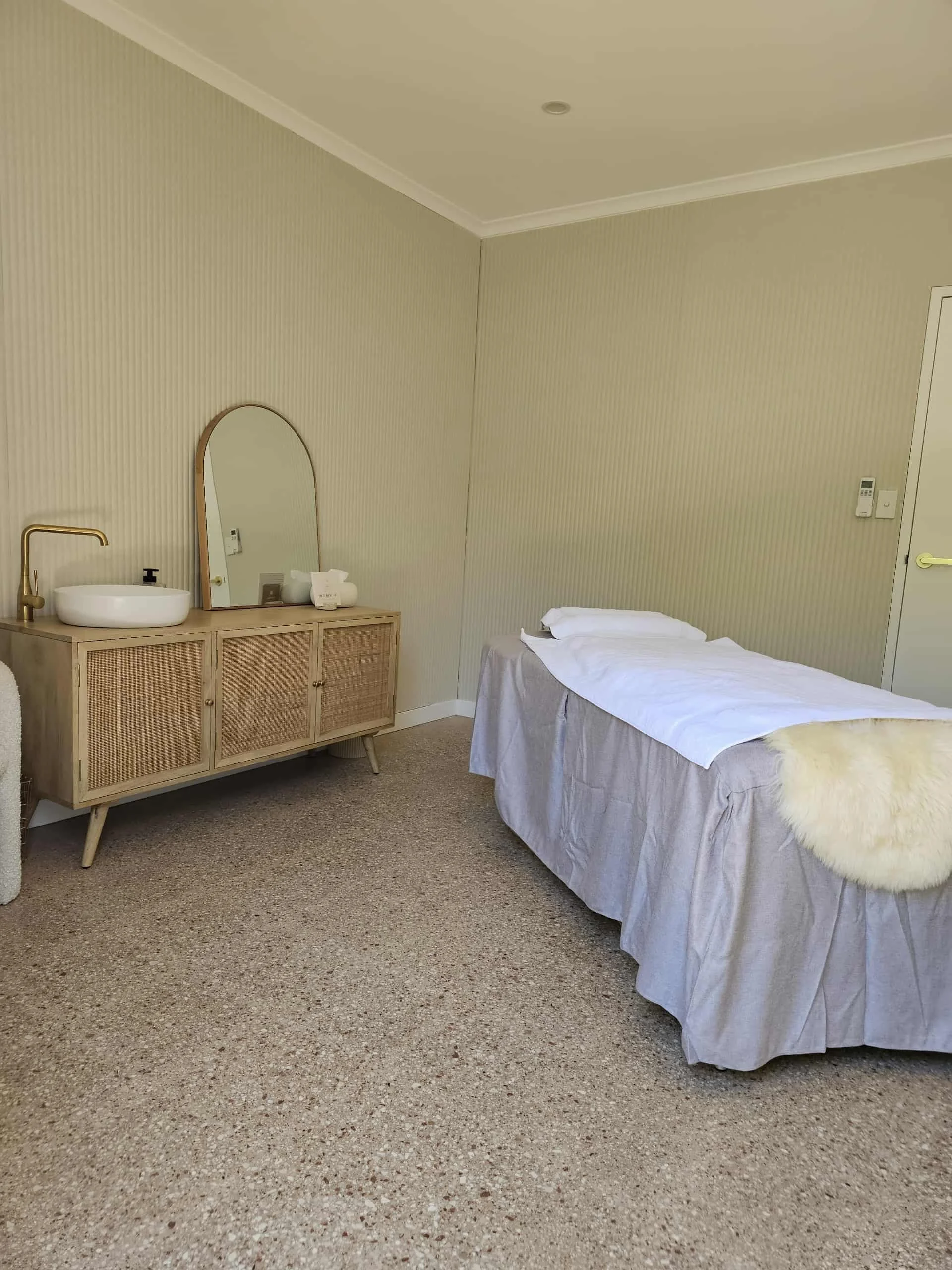 A minimally decorated bedroom with a single bed made with white linens, a fluffy cream duvet at the foot, and a pillow. There is a wooden cabinet with a mirror, a sink with a gold faucet, and some toiletries on top, along with a soap dispenser. The r