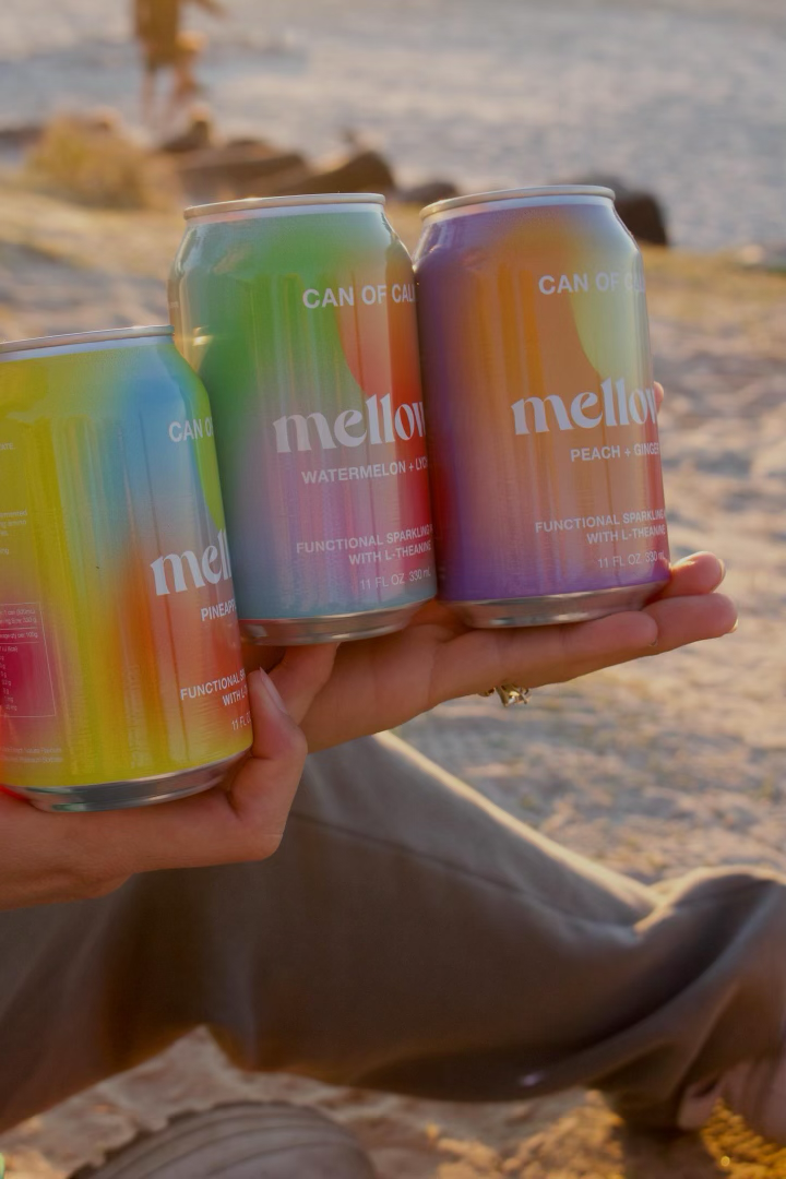 Three cans of Mellow melon, watermelon & uva, and peach & ginger flavored sparkling beverages being held by a person near a beach at sunset.
