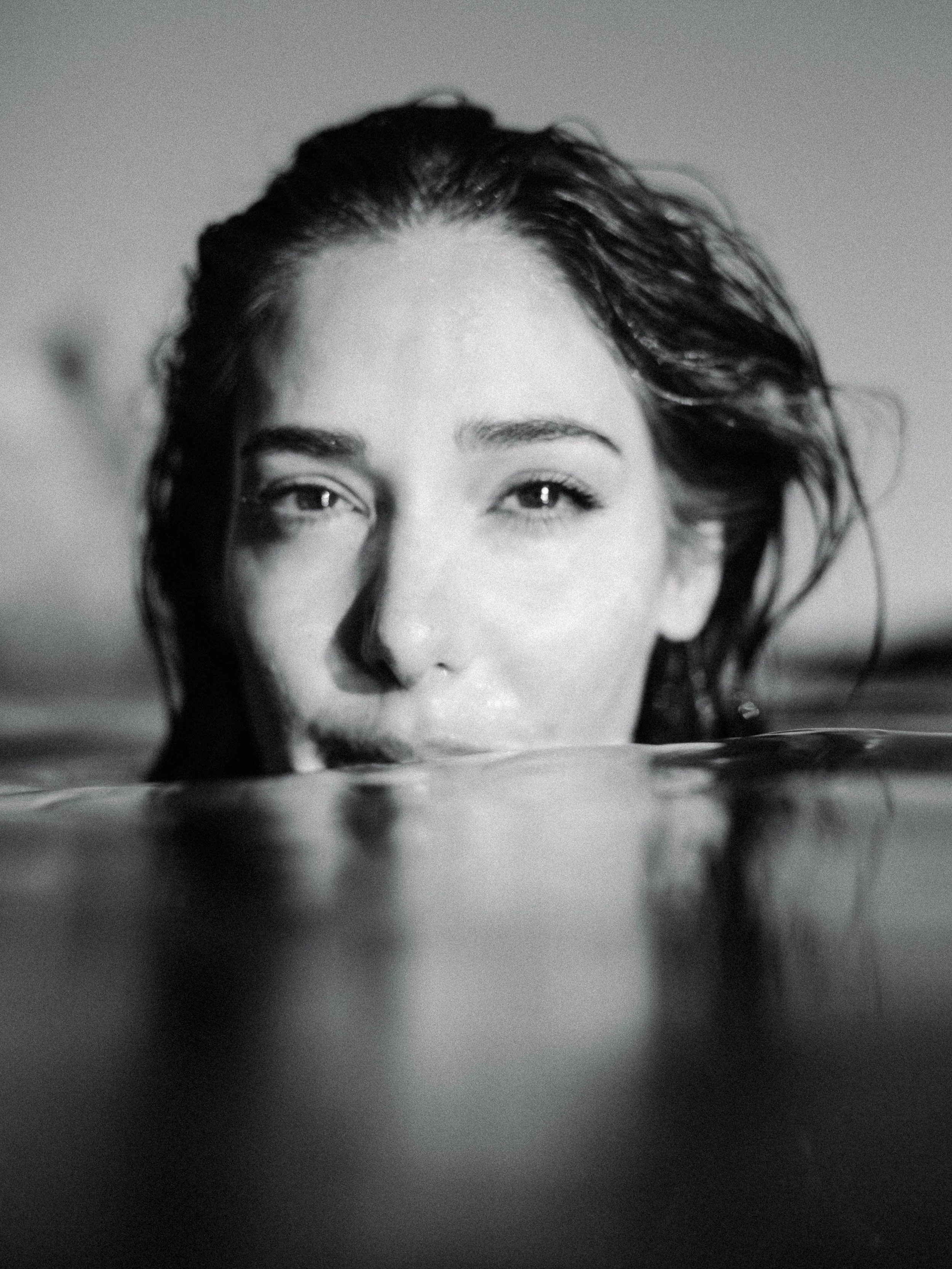 A black and white close-up shot of a woman with her body and the lower portion of her face, mostly her chin, dipped below the surface. There's a beautiful catchlight reflected in her eyes.