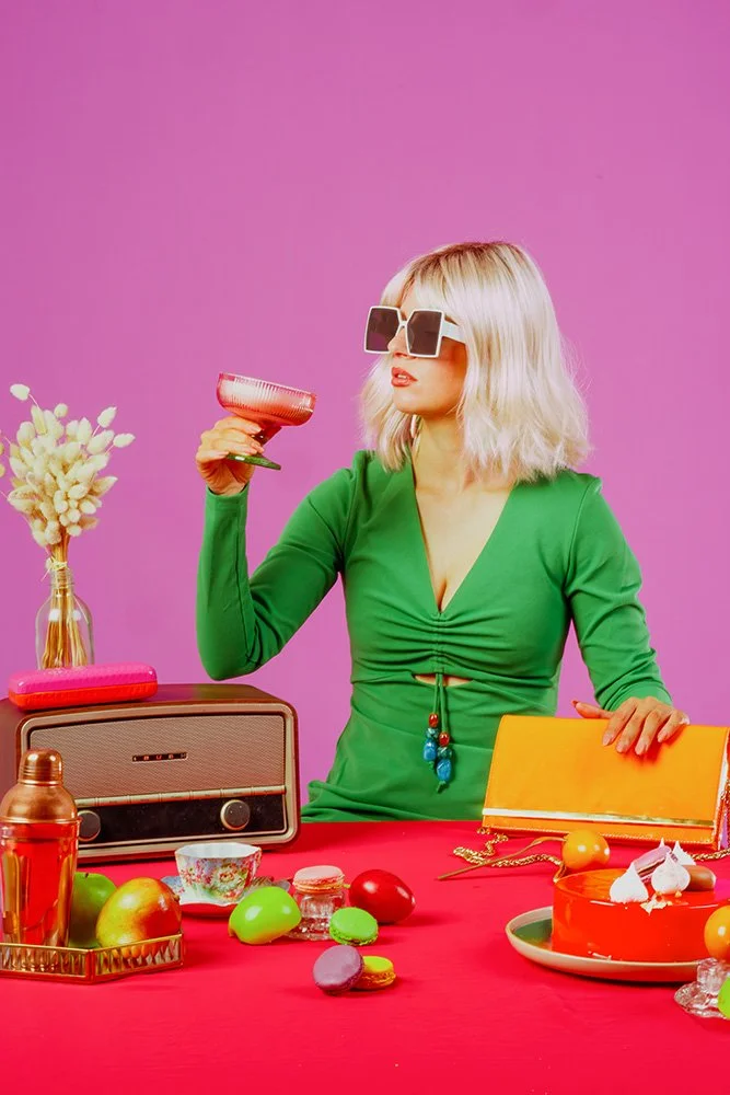 A blonde woman is wearing a green dress and holding a cocktail. She is surrounded by bold, bright colours, with sweets and fruit placed on the table in front of her. A vintage radio adds to the indoor setting, which features a pink wall and red table