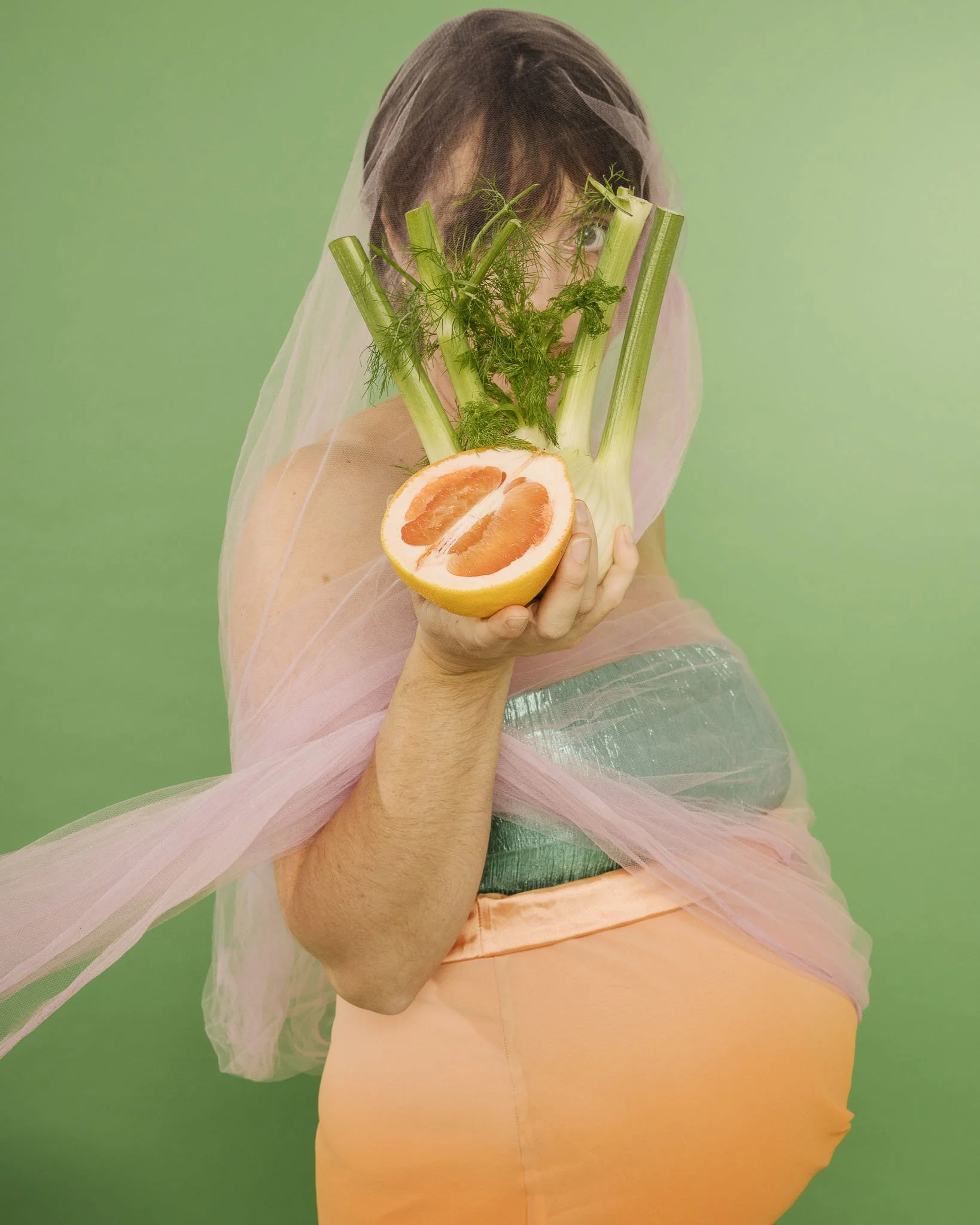 A pregnant woman holds a fennel bulb and half a grapefruit in front of her face. She has cropped dark hair and is dressed in a sparkly cropped tube top paired with a long orange skirt.
