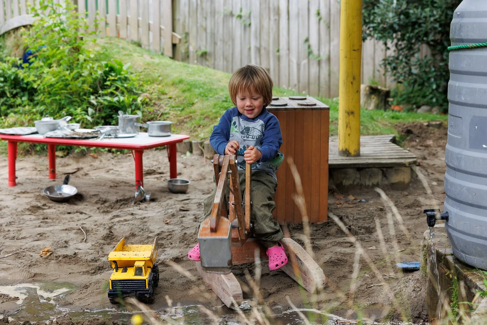 A young child playing outdoors on a sandbox with toy construction equipment, surrounded by toy bowls and utensils, with greenery and a wooden fence in the background.