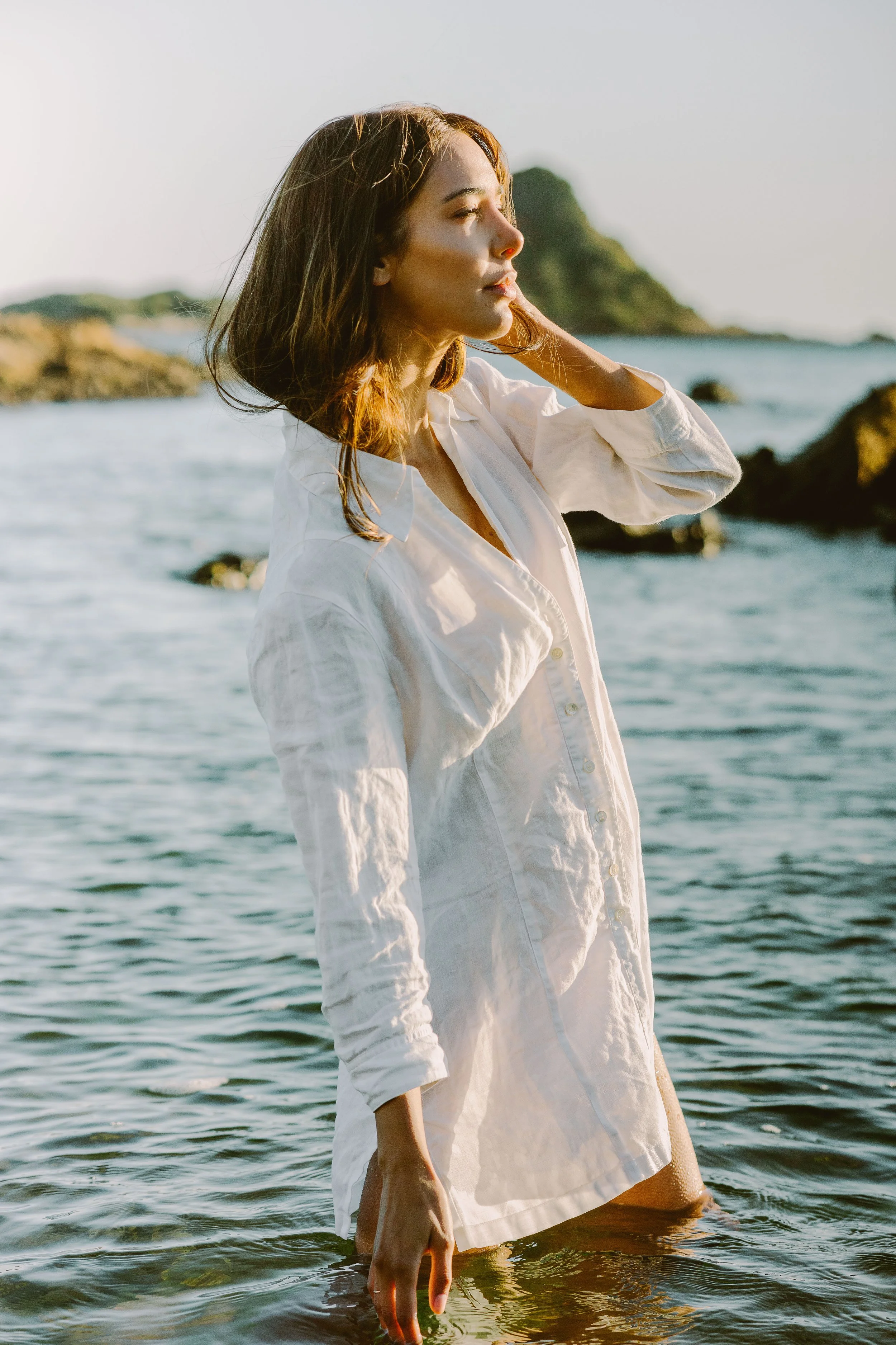 A woman is standing in the ocean, wearing a white shirt. Her long dark hair is tousled softly over her shoulders as she looks off-camera to her right. The scene captures a fashion photo shoot vibe with elements of natural beauty and light.
