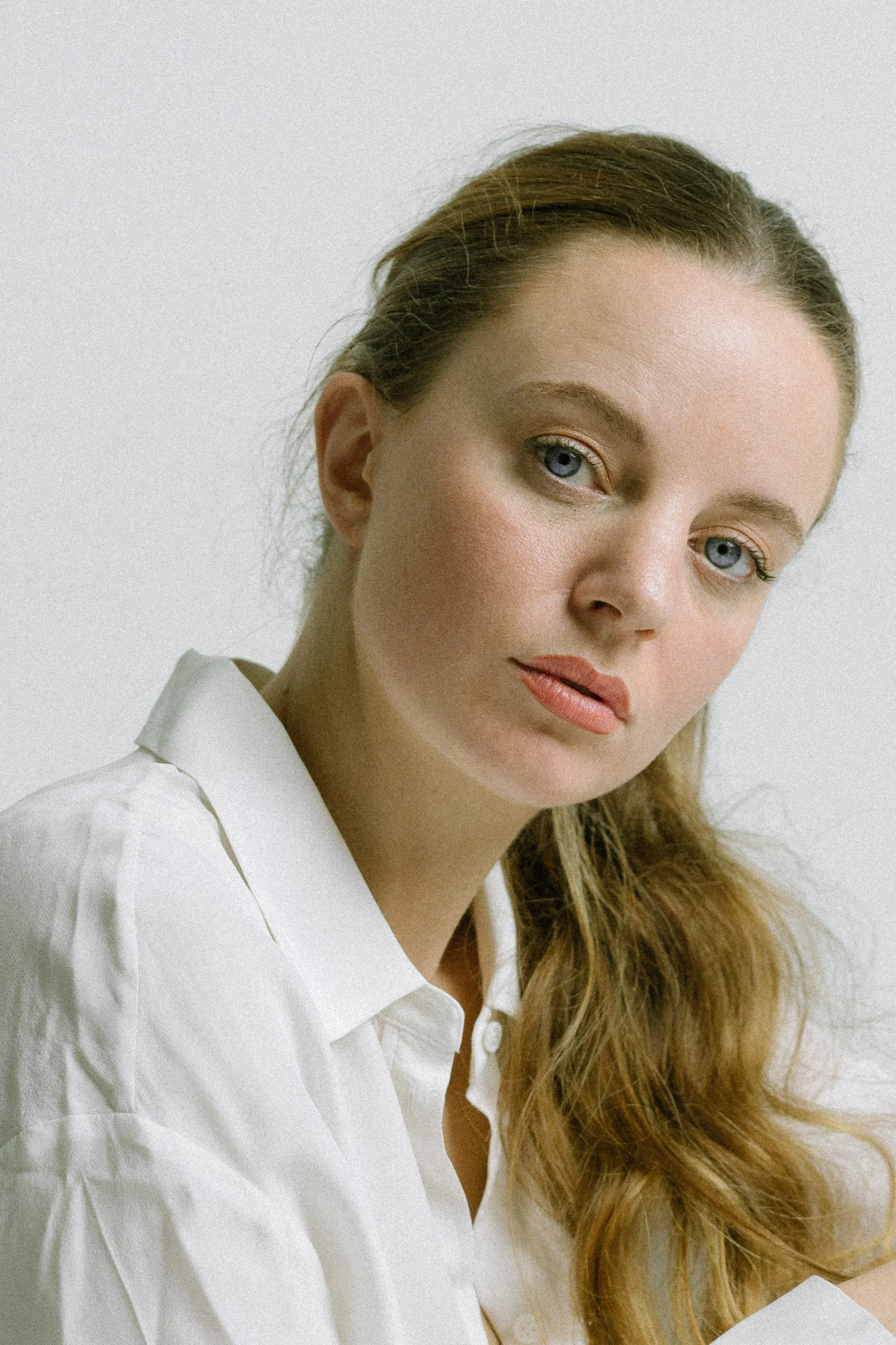 A close-up studio portrait of a woman against a white backdrop. She is dressed in a loose white shirt and her long, medium brown hair is tied into a ponytail and draped over one shoulder. She looks directly at the camera with a calm gaze.