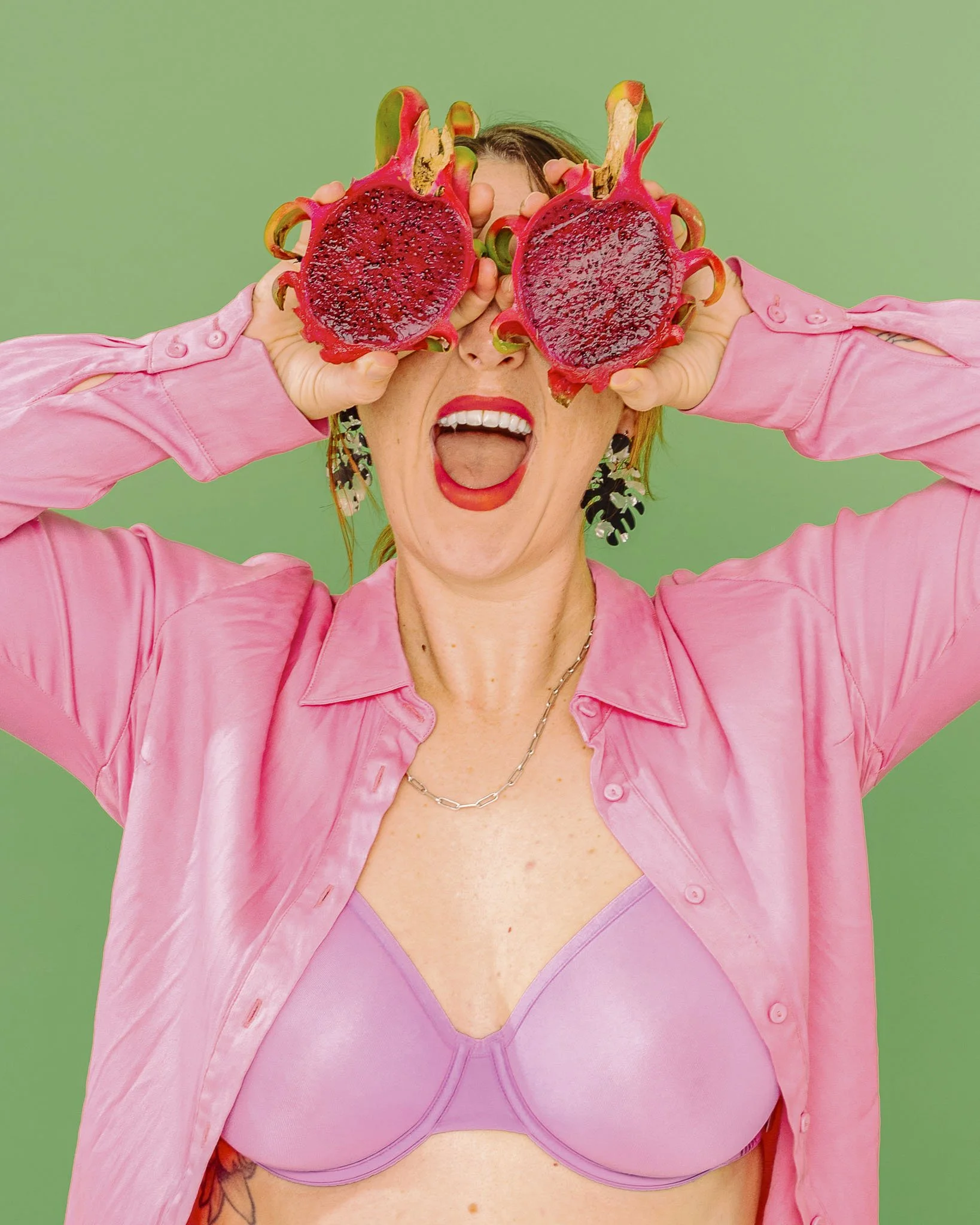 A woman is playfully holding a dragon fruit in front of her eyes, concealing her gaze. She has a big, open-mouthed smile. The vibrant pink colours and her clothing contrast with the vibrant green backdrop, adding to the playful vibe of the image.