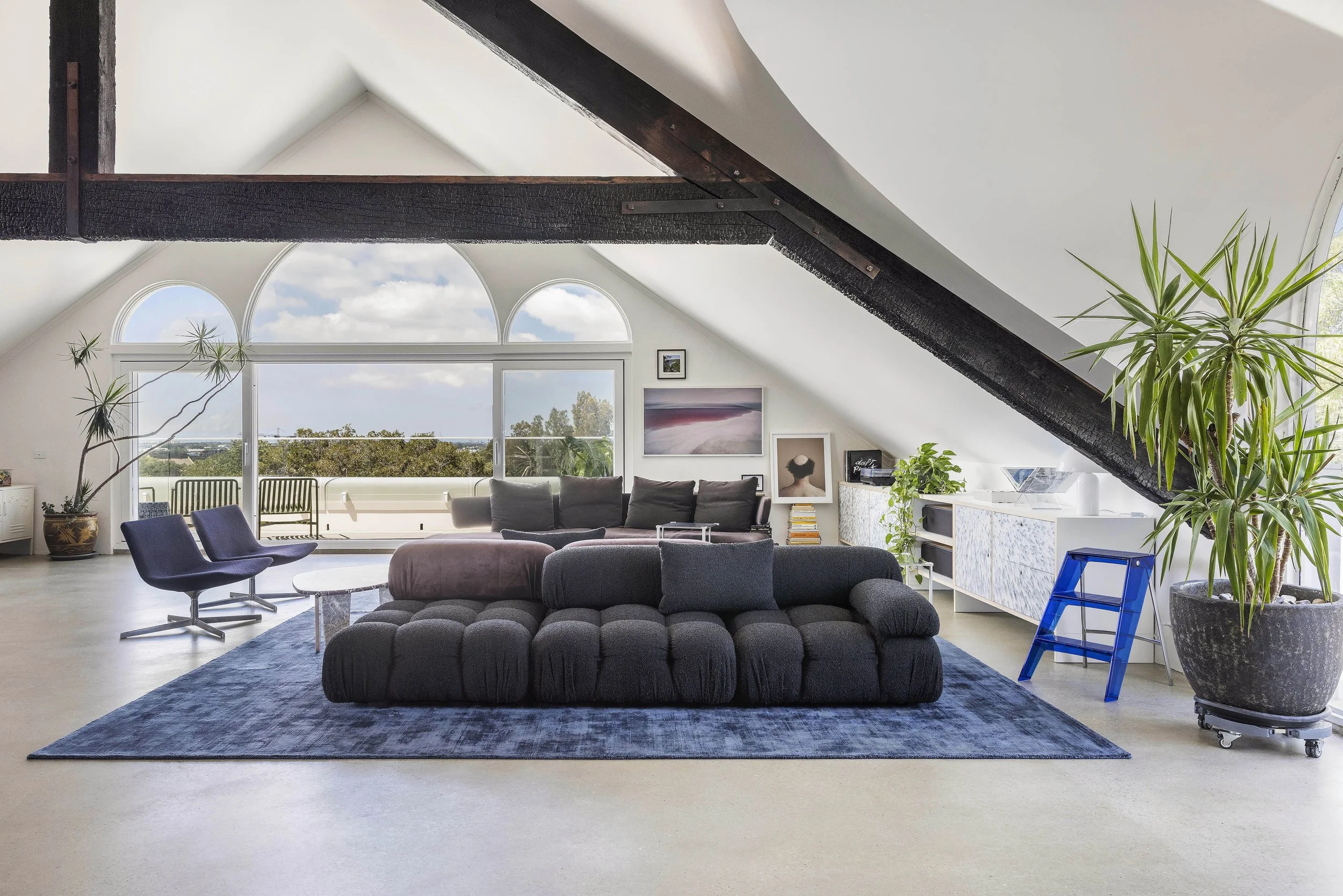 Modern living room in an attic with sloped ceiling, large window with balcony, black sofa, armchair, potted plants, paintings on wall, contemporary furniture, blue rug, and natural light.