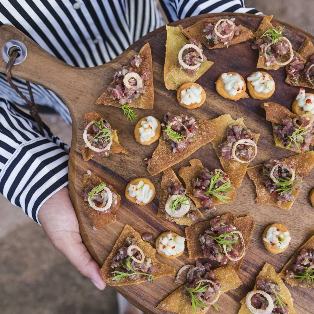 A wooden serving board with assorted gourmet appetizers, including crispy chips topped with meat and herbs, and small round crackers with cream cheese and herbs. Person holding the board is wearing a striped shirt.