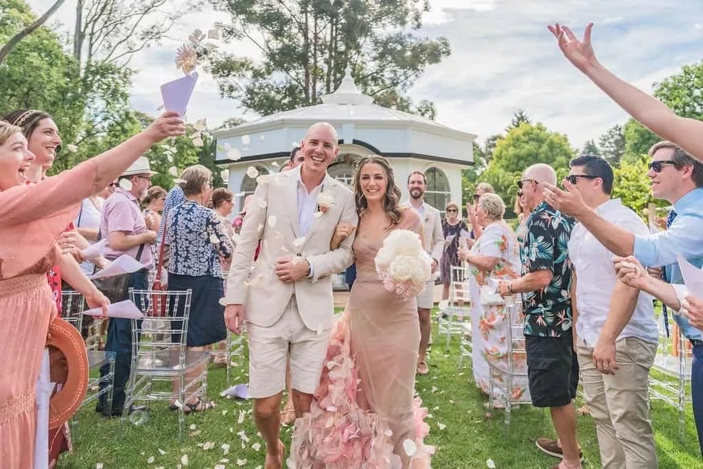 A bride and groom walking down an aisle outdoors, surrounded by cheering guests tossing flower petals, during a wedding ceremony with green trees and a white gazebo in the background.