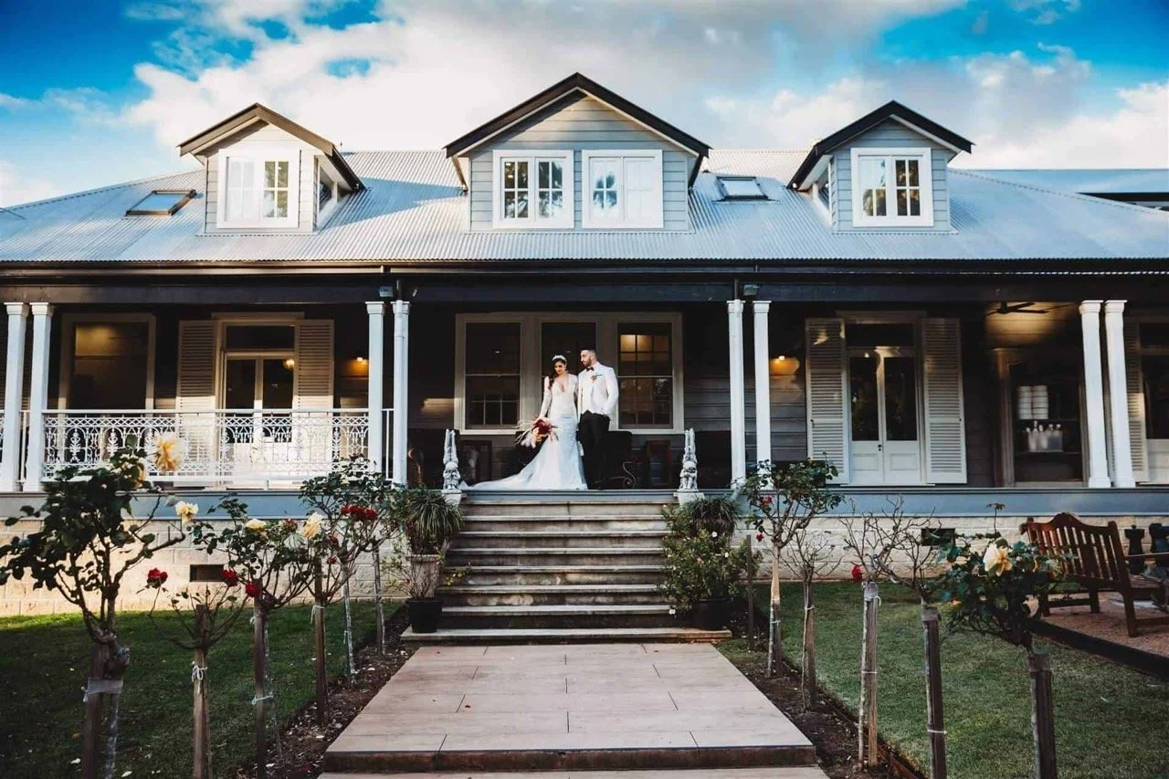 Bride and groom standing on the steps of a traditional house with a veranda, surrounded by a garden with roses.