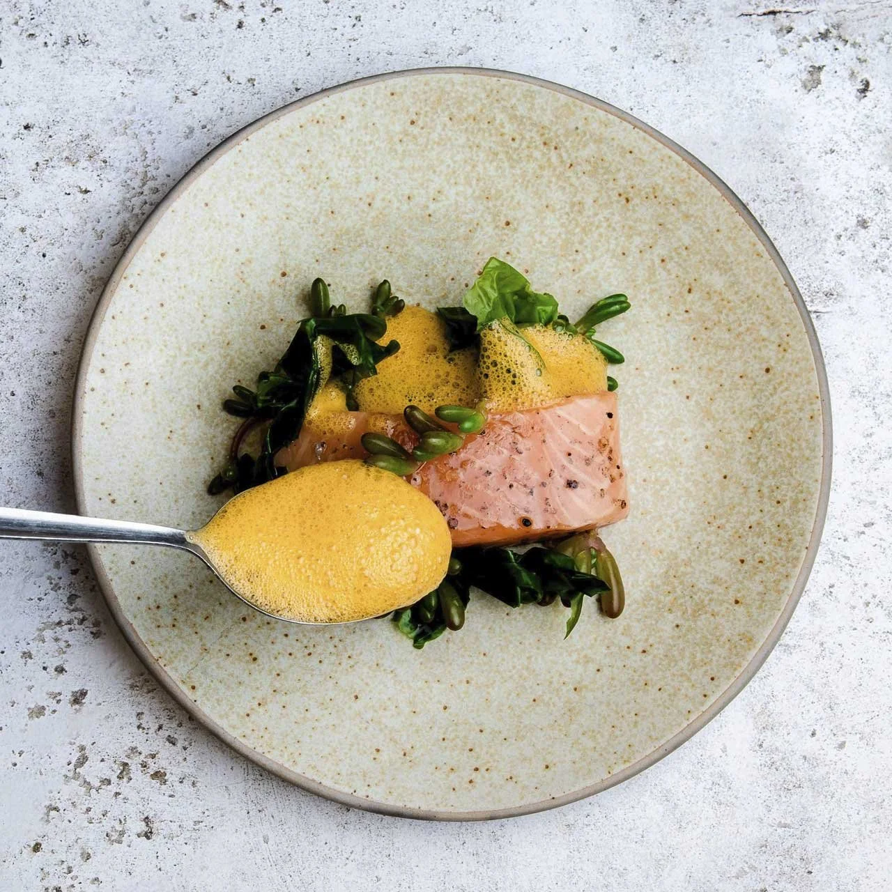 Plate with salmon fillet, leafy greens, yellow foam sauce, spoon adding more foam. Gourmet food presentation.