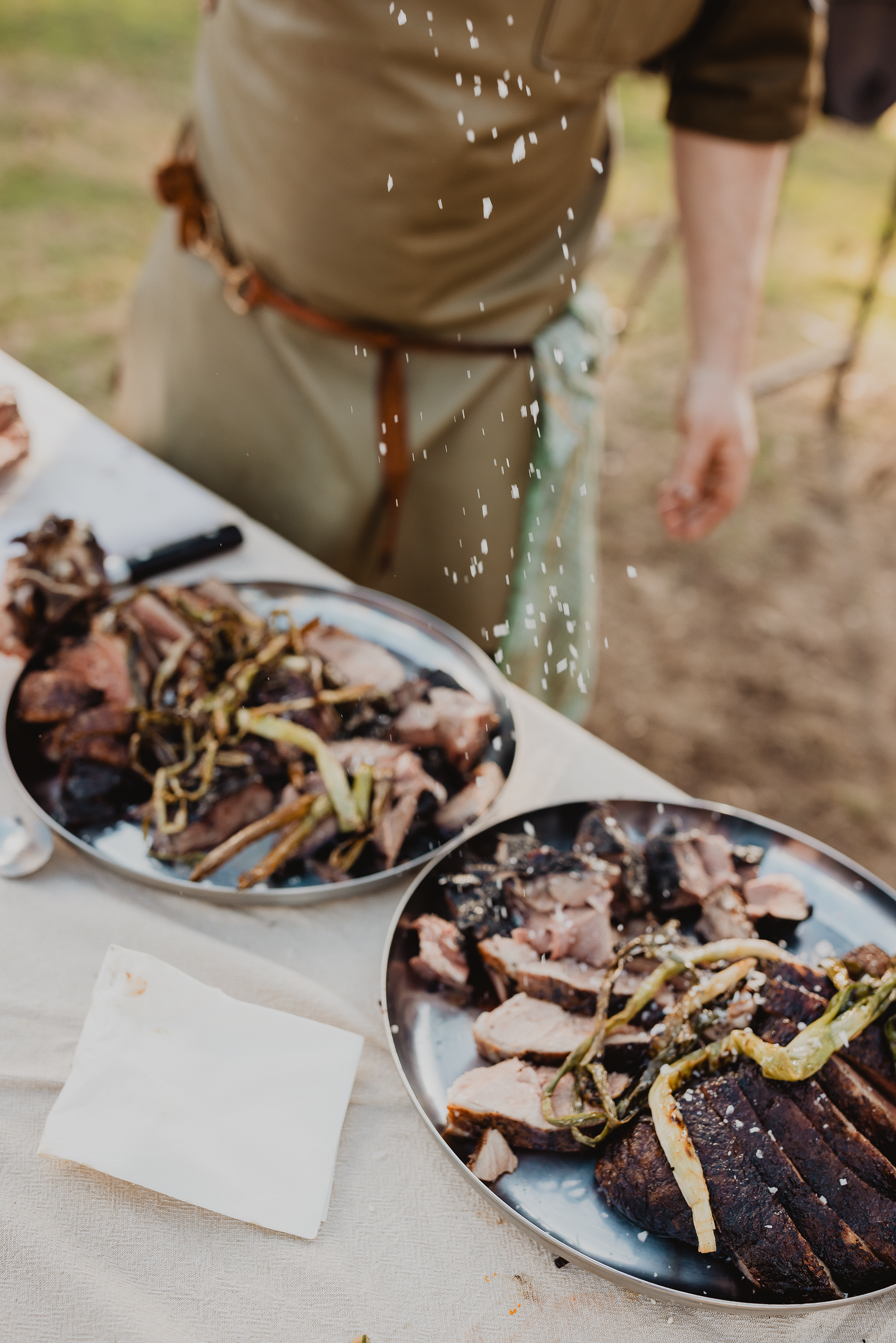 Person seasoning cooked meat and vegetables on plates outdoors.