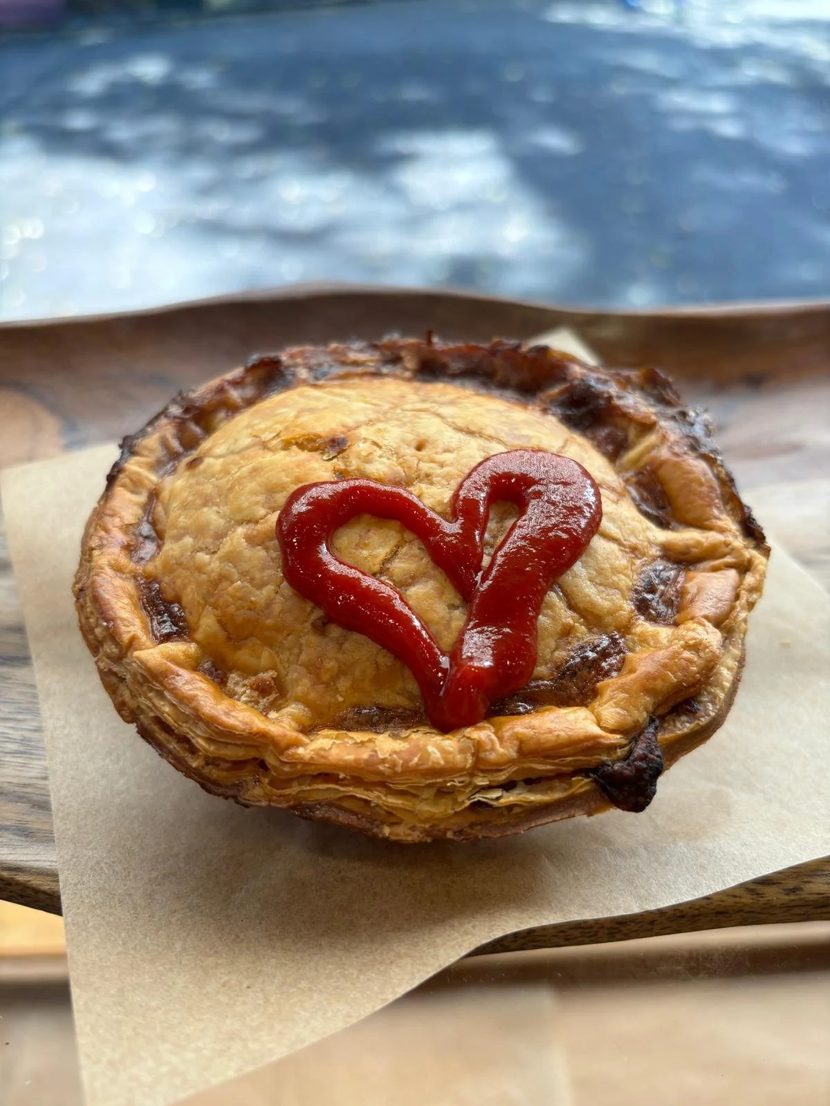 A meat pie topped with a heart-shaped design made of ketchup, served on a piece of parchment paper on a wooden tray.