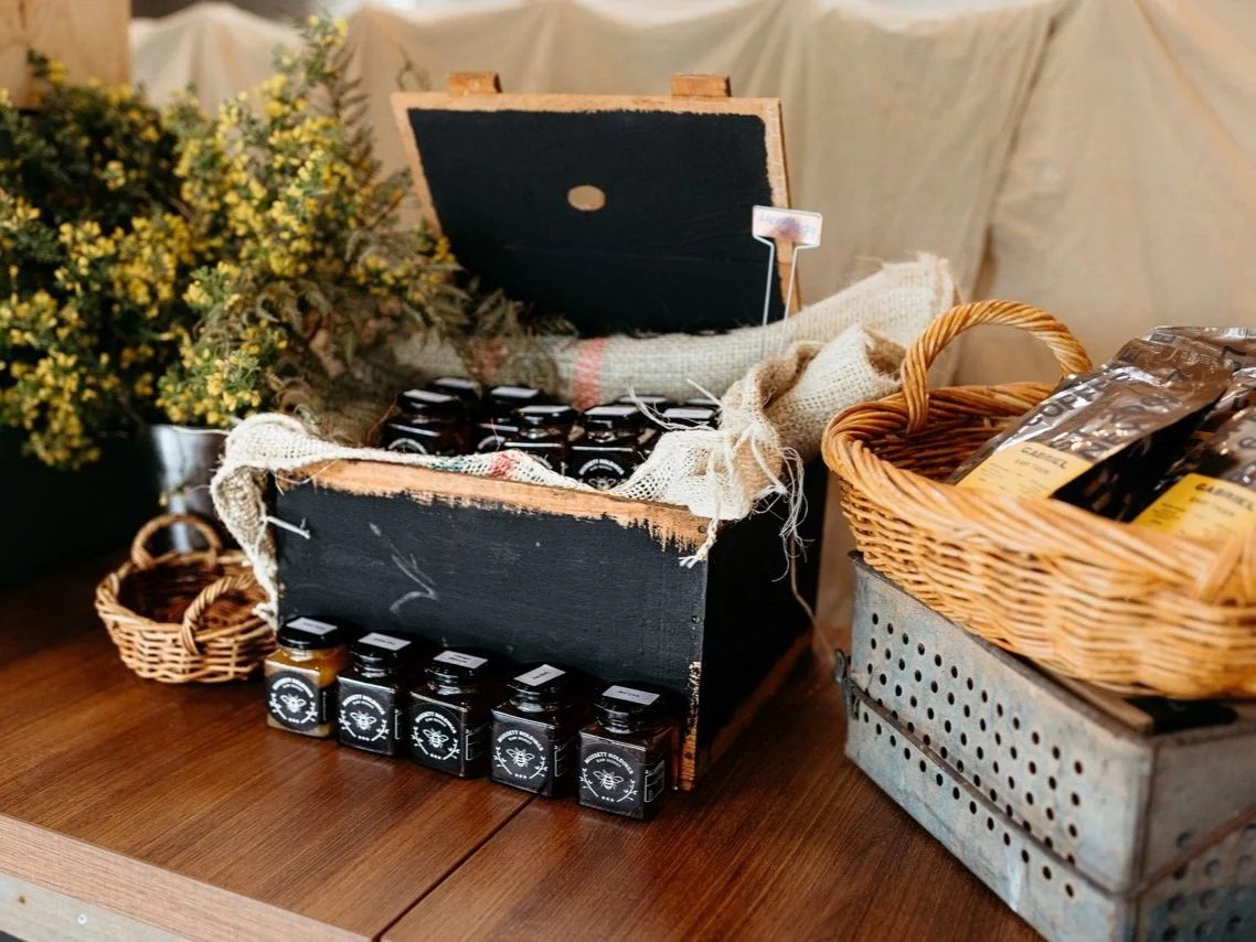 Display of various jars and packaged goods on a wooden table, with decorative greenery and wicker baskets, suggesting a rustic market or shop setting.