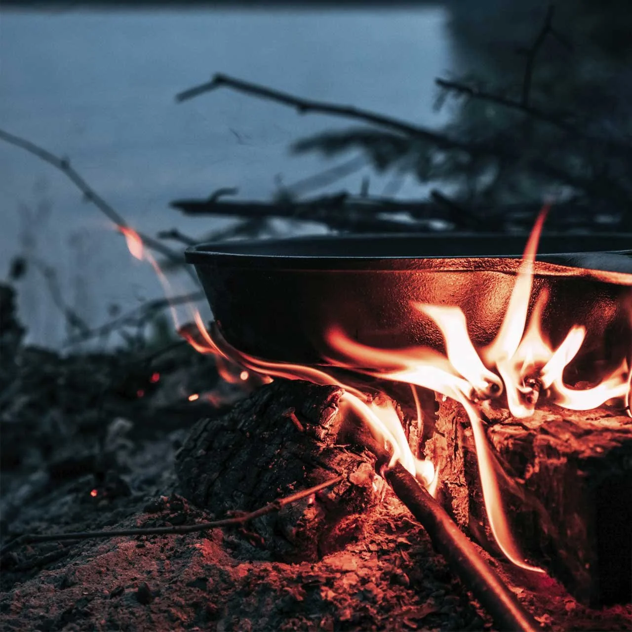 Cast iron skillet on an open fire with flames and logs in an outdoor setting.