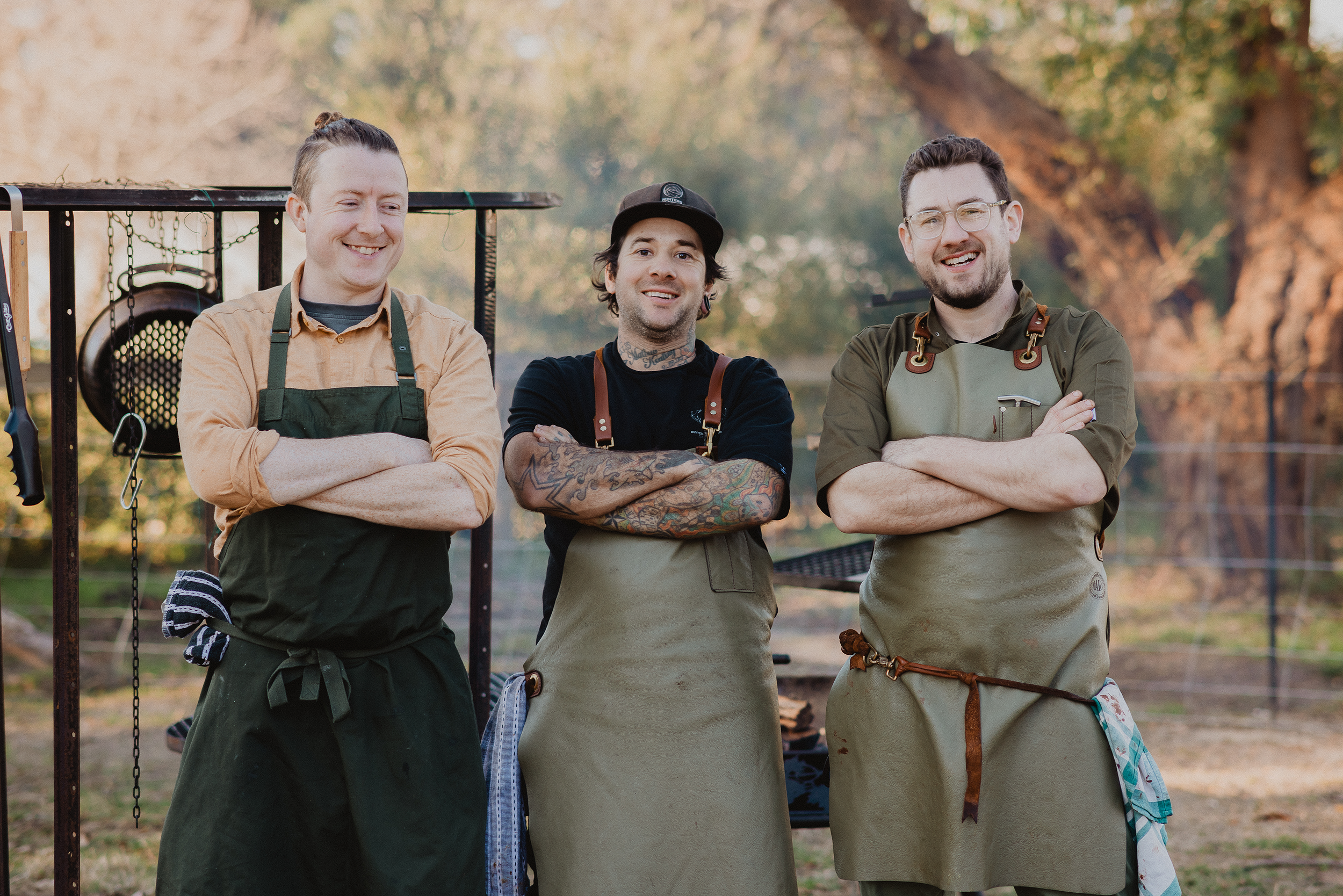 Three men wearing aprons standing outdoors with arms crossed, smiling. Cooking utensils visible in the background.