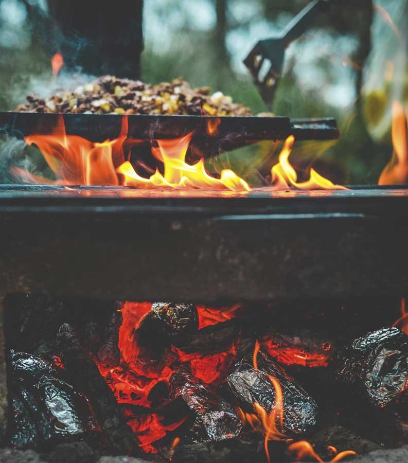 Outdoor grill with flames and food cooking on the griddle, with glowing charcoal below.