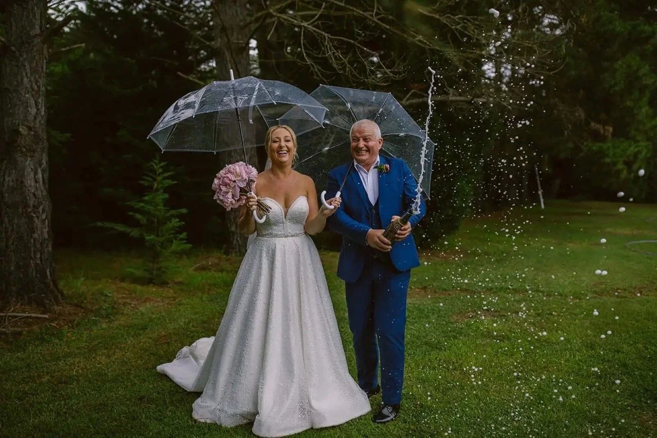 A bride and groom in a forest setting, holding clear umbrellas and celebrating with a champagne bottle. The bride is in a white dress holding a bouquet, and the groom is in a blue suit. Champagne is visibly spraying.