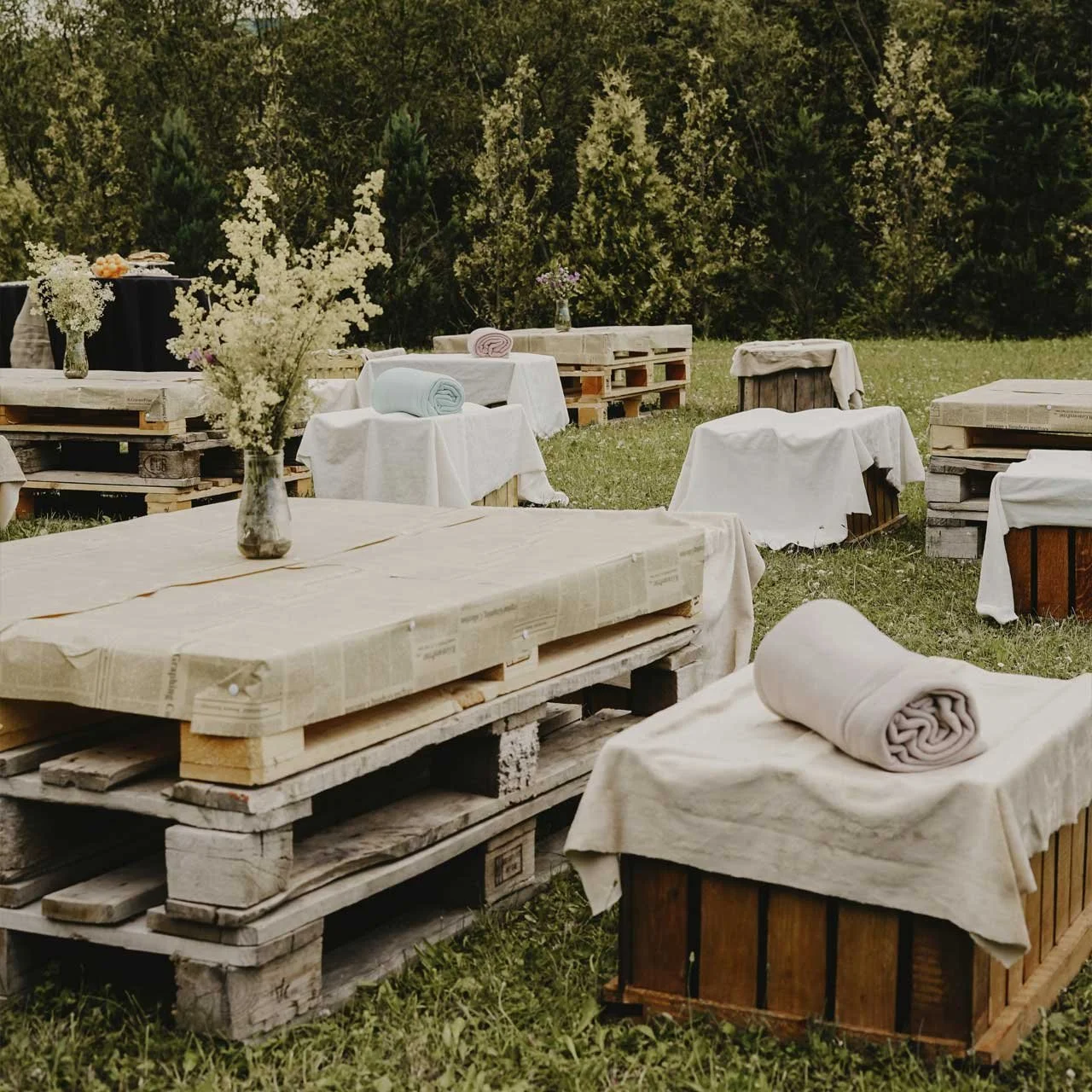 Outdoor rustic seating arrangement with wooden pallet tables covered in cloth and decorated with vases of flowers; rolled blankets placed on wooden crates; trees and grass in the background.