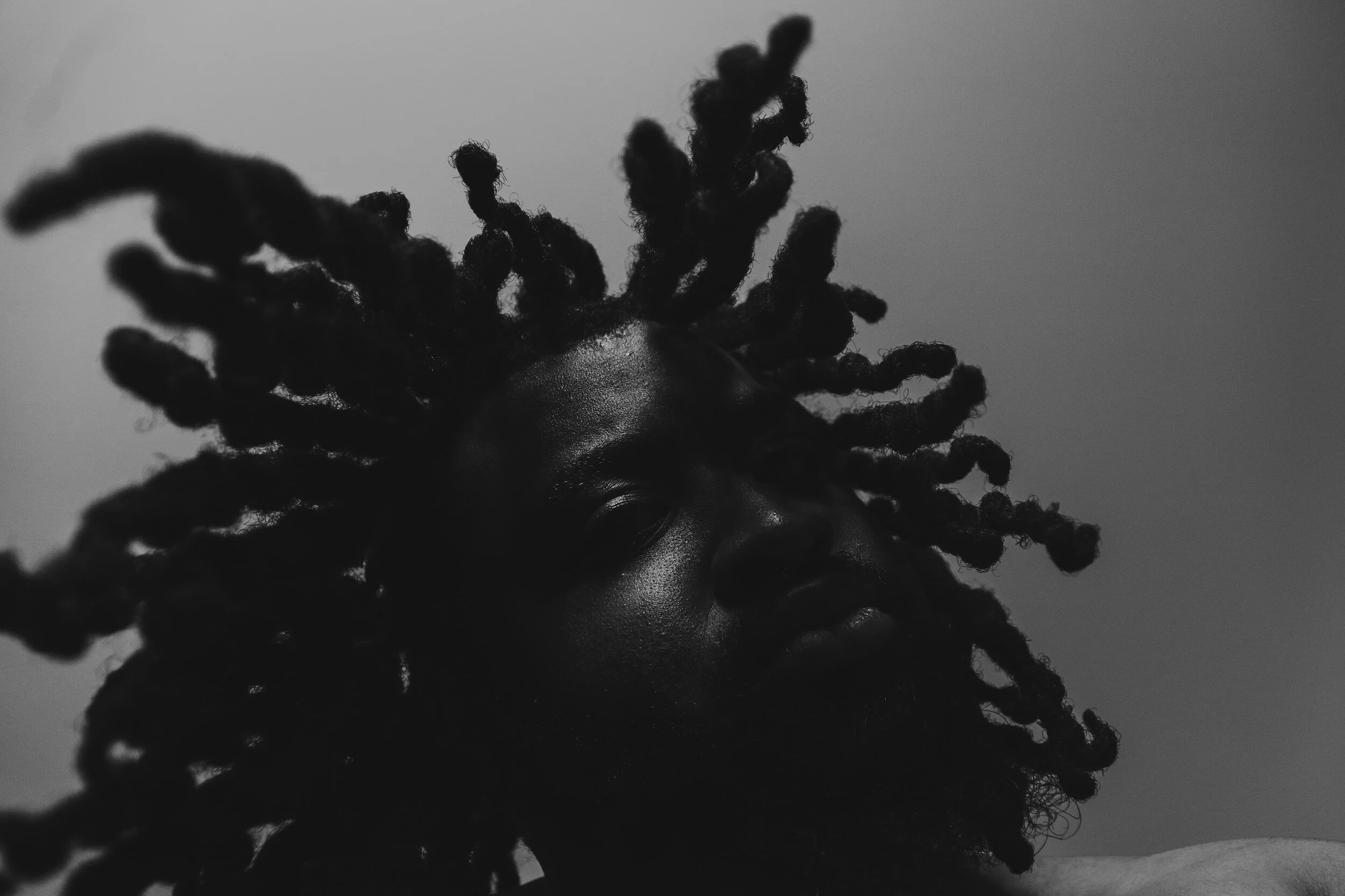 Close-up of a person with thick dreadlocks, partially shaved head, dark skin, and a serious expression, in black and white.