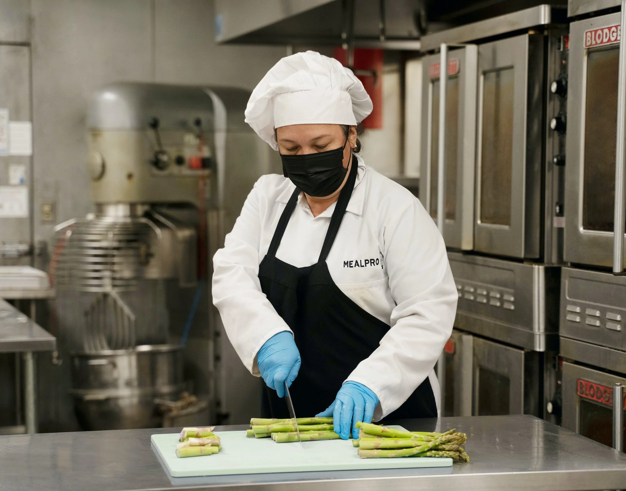 A chef wearing a black mask and blue gloves slices fresh asparagus on a cutting board in a commercial kitchen.