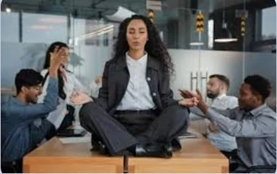 A woman sitting cross-legged on a table in a meeting room, with colleagues around her, some holding phones, in a professional office setting.