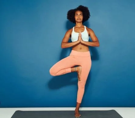 Woman practicing yoga in a tree pose with hands in prayer position in front of a blue wall.